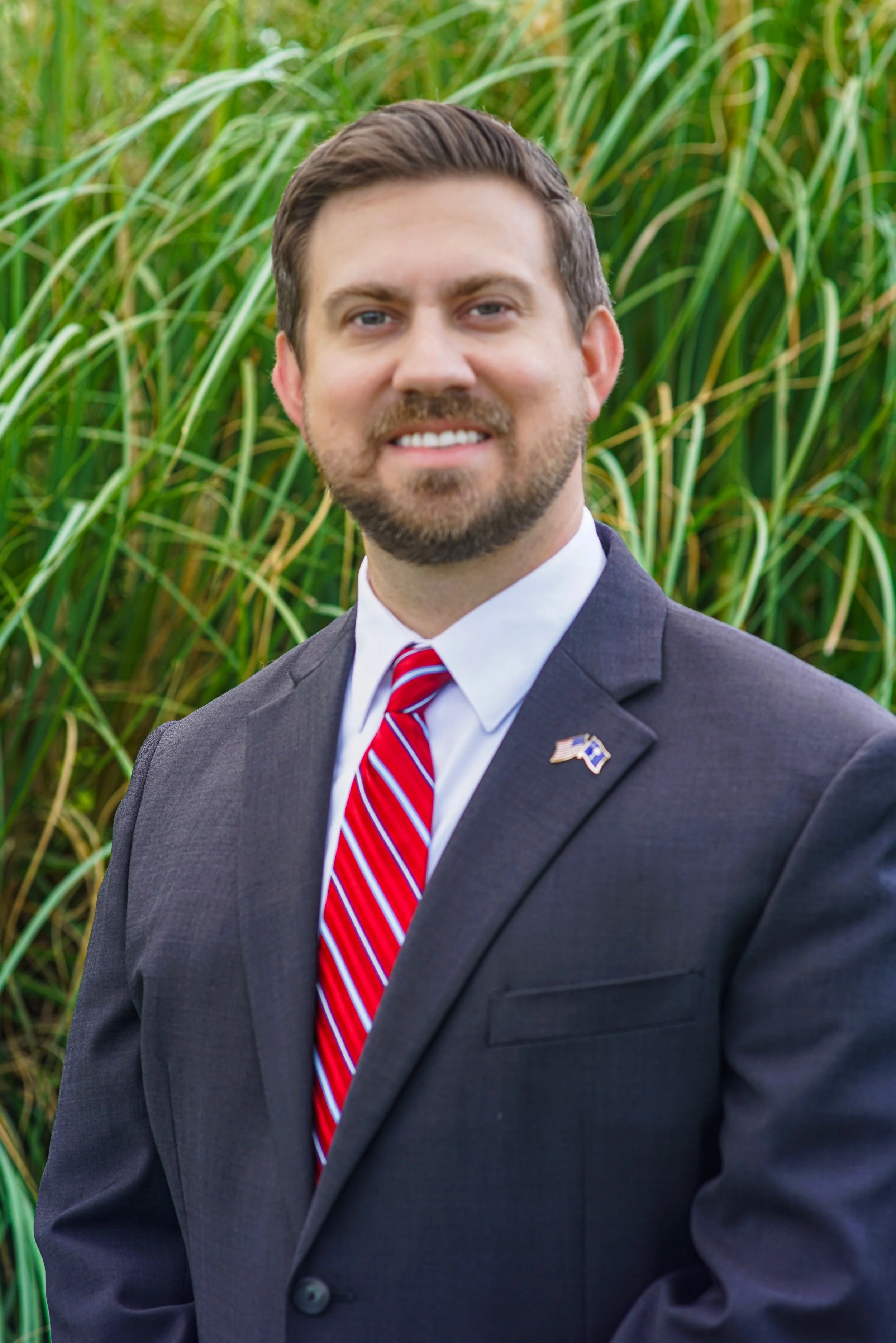 A man in a dark suit and red and white striped tie standing outdoors in front of green plants, with an American flag lapel pin on his jacket.