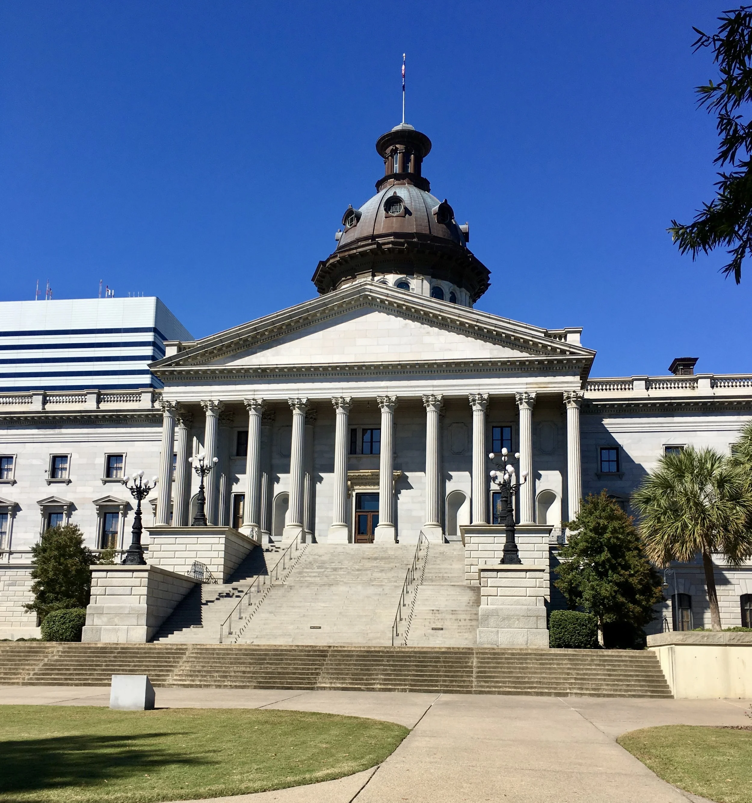 A neoclassical government building with white stone, columns, steps, and a central dome, set against a clear blue sky with some trees in the foreground.