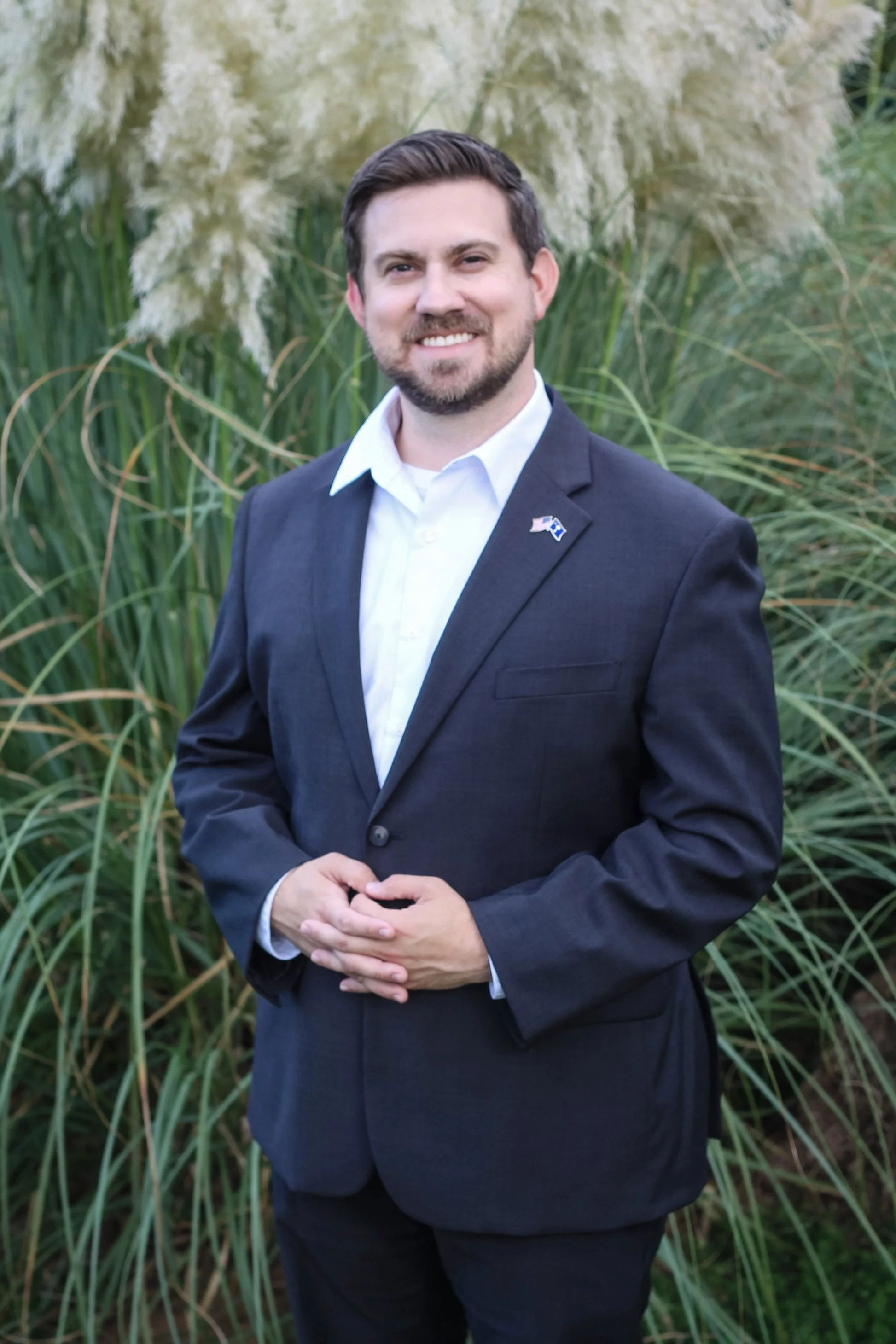 A man in a dark suit and white shirt standing outdoors in front of tall grasses, smiling at the camera with hands clasped.