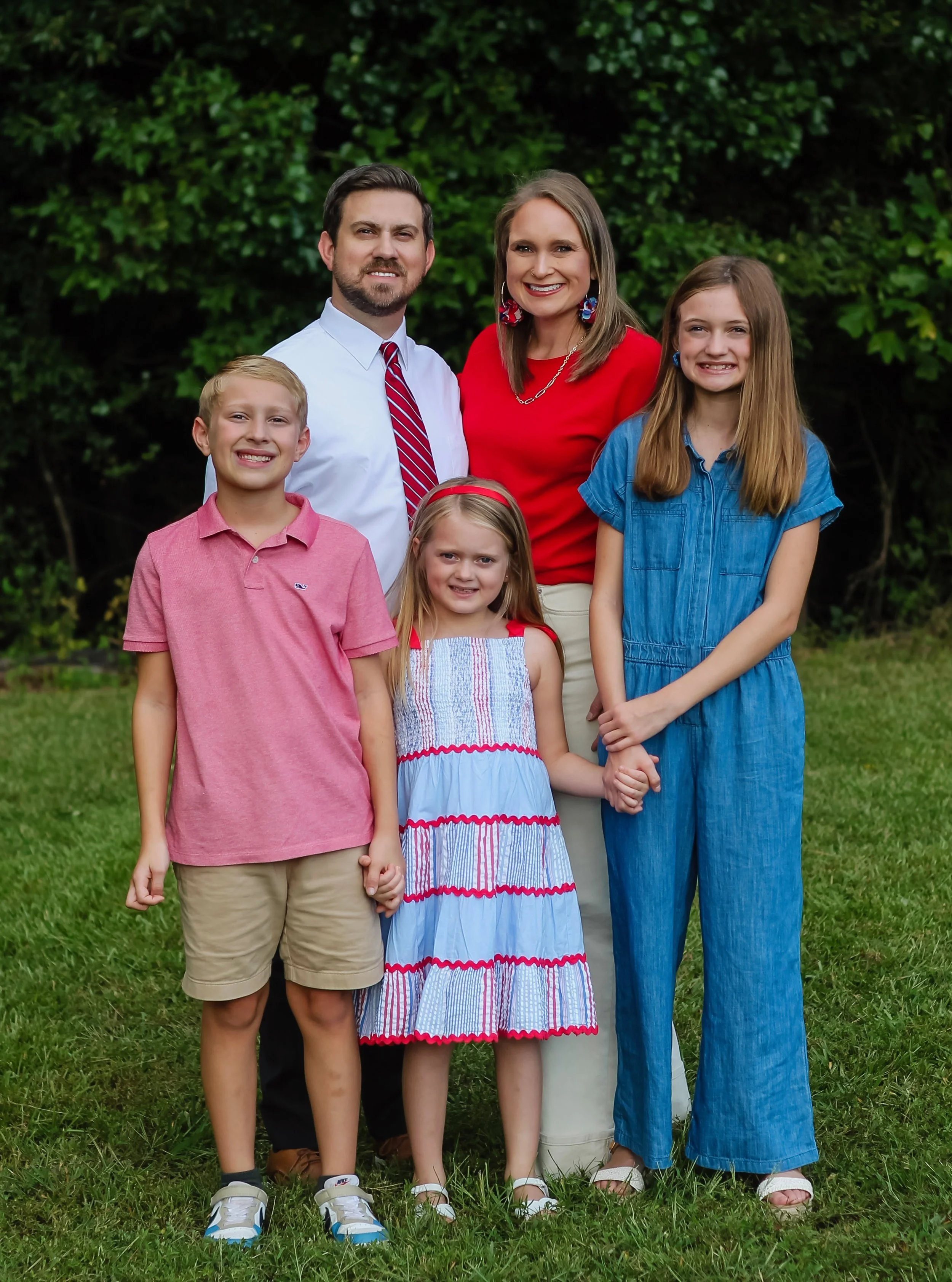 Family of six posing outdoors in front of green trees, smiling and holding hands, dressed in casual summer clothing.
