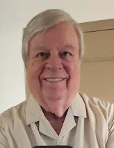 A smiling older man with gray hair wearing a light-colored shirt.