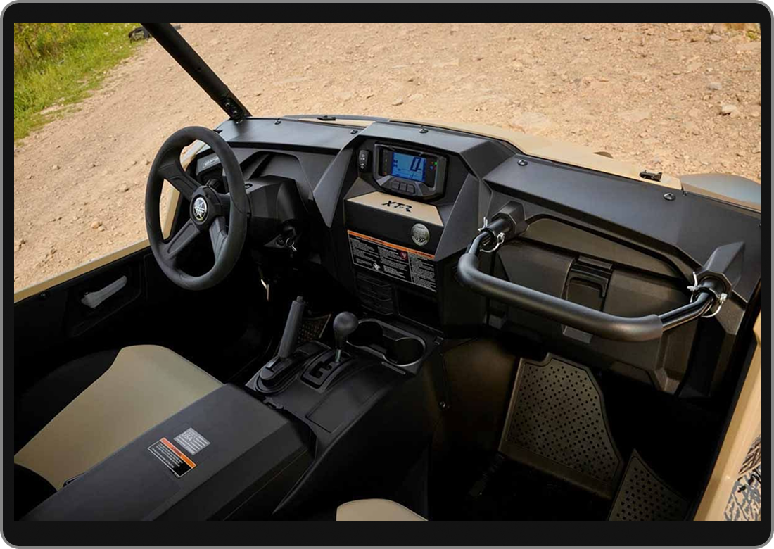 Interior view of a utility vehicle with a steering wheel, digital display, gear shifter, and safety bar on a dirt road.