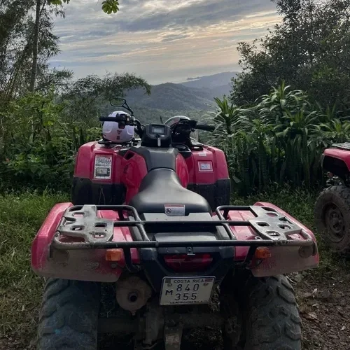 Red all-terrain vehicle (ATV) parked on a dirt path near greenery with hills and a cloudy sky in the background.