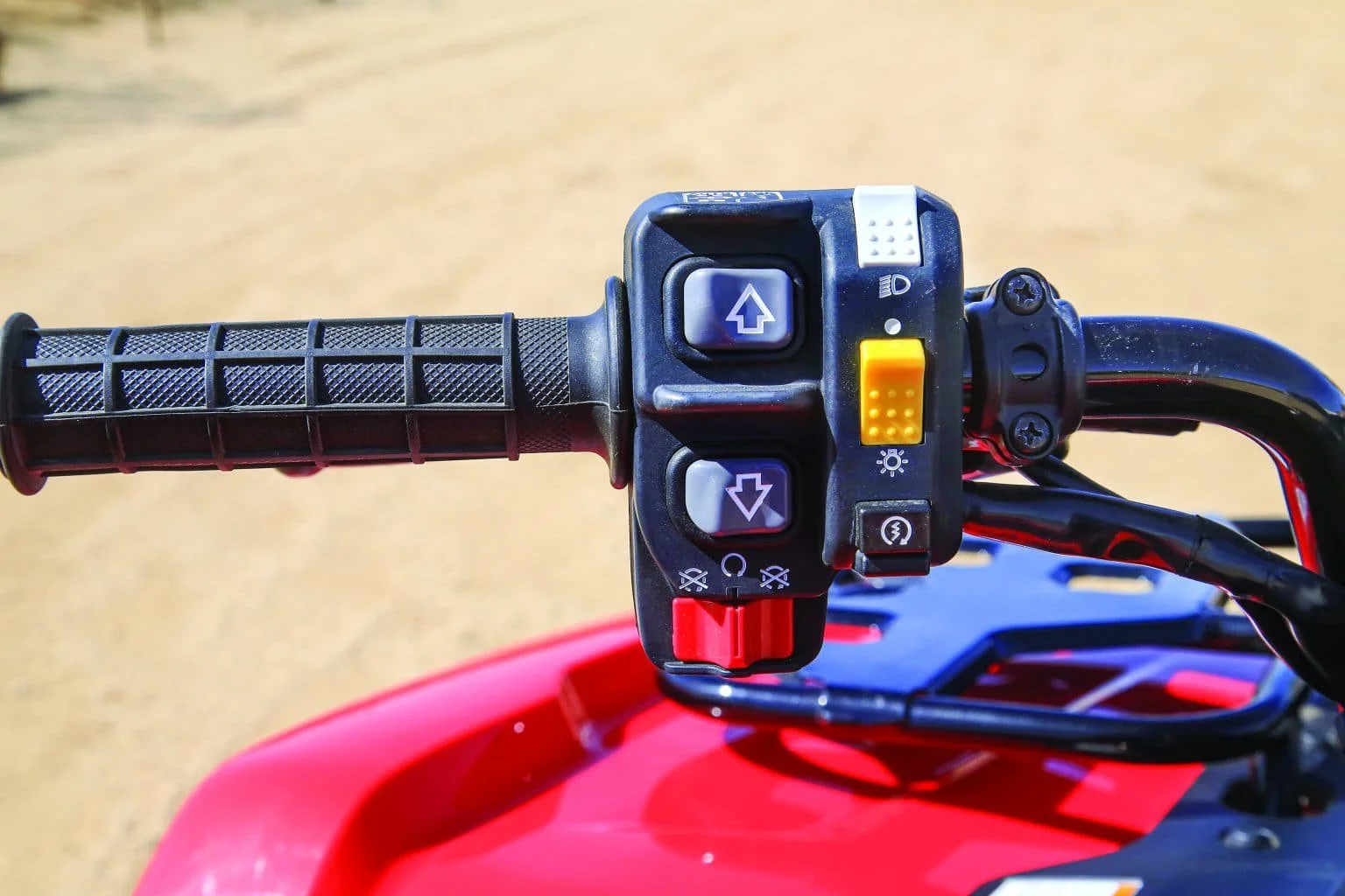 Close-up of right handlebar controls on a red ATV side, showing a textured grip and various buttons for navigation and light control, with a dirt ground background.