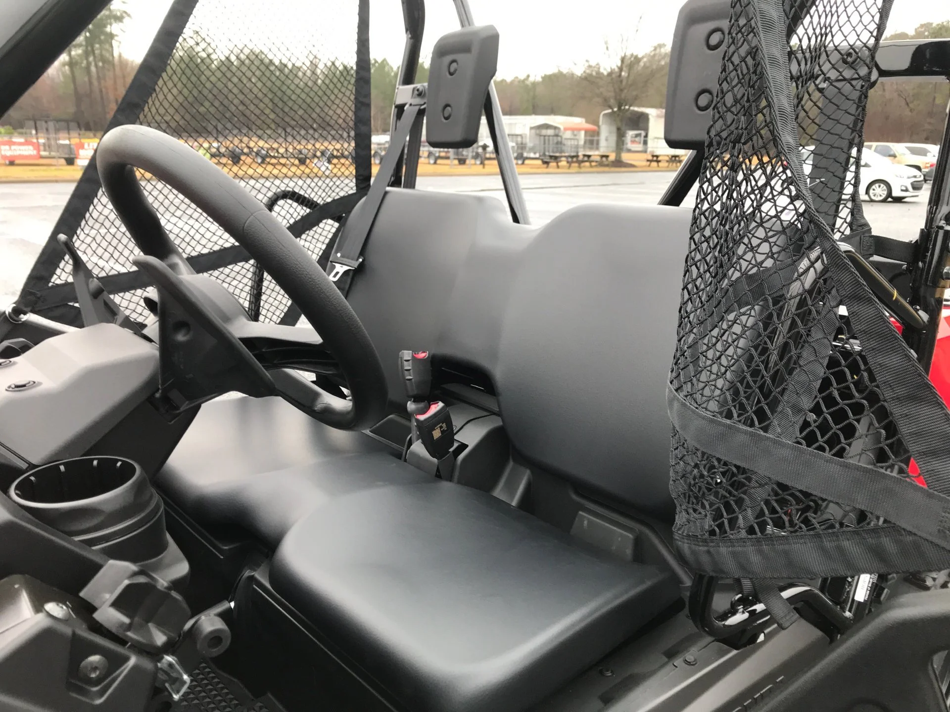 Interior view of a utility vehicle or ATV showing a black steering wheel, a cup holder, a gray dashboard, and a mesh storage pocket on the right side, with a parking lot and trees visible through the windows.