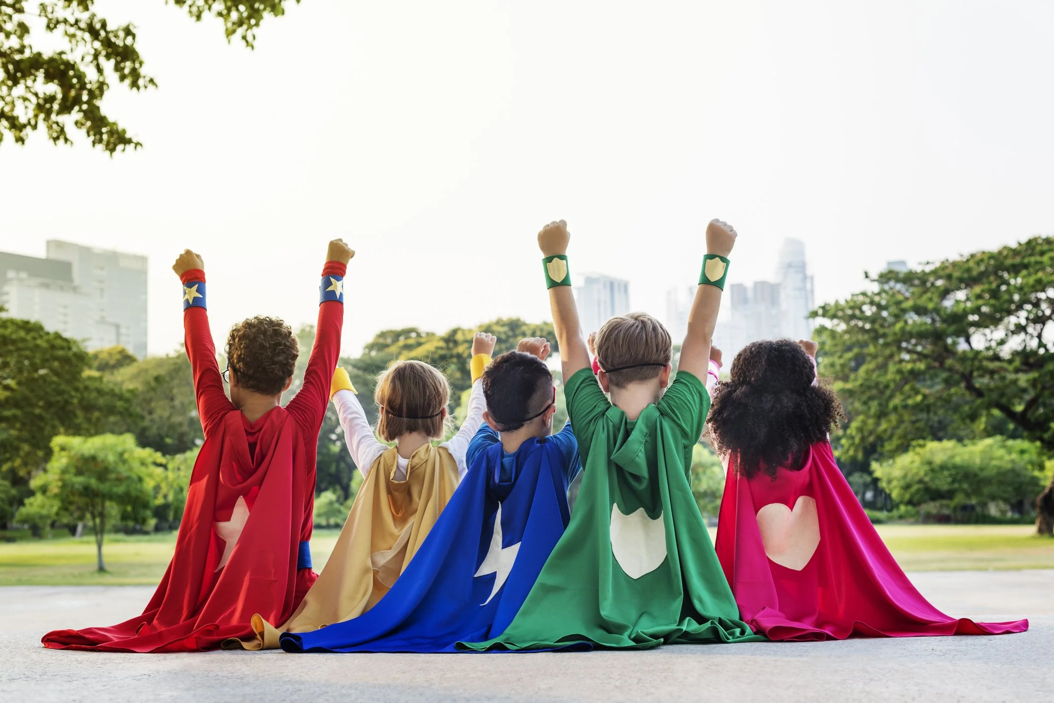Five children in colorful superhero capes sit side by side with their backs to the camera, fists raised, in a park.