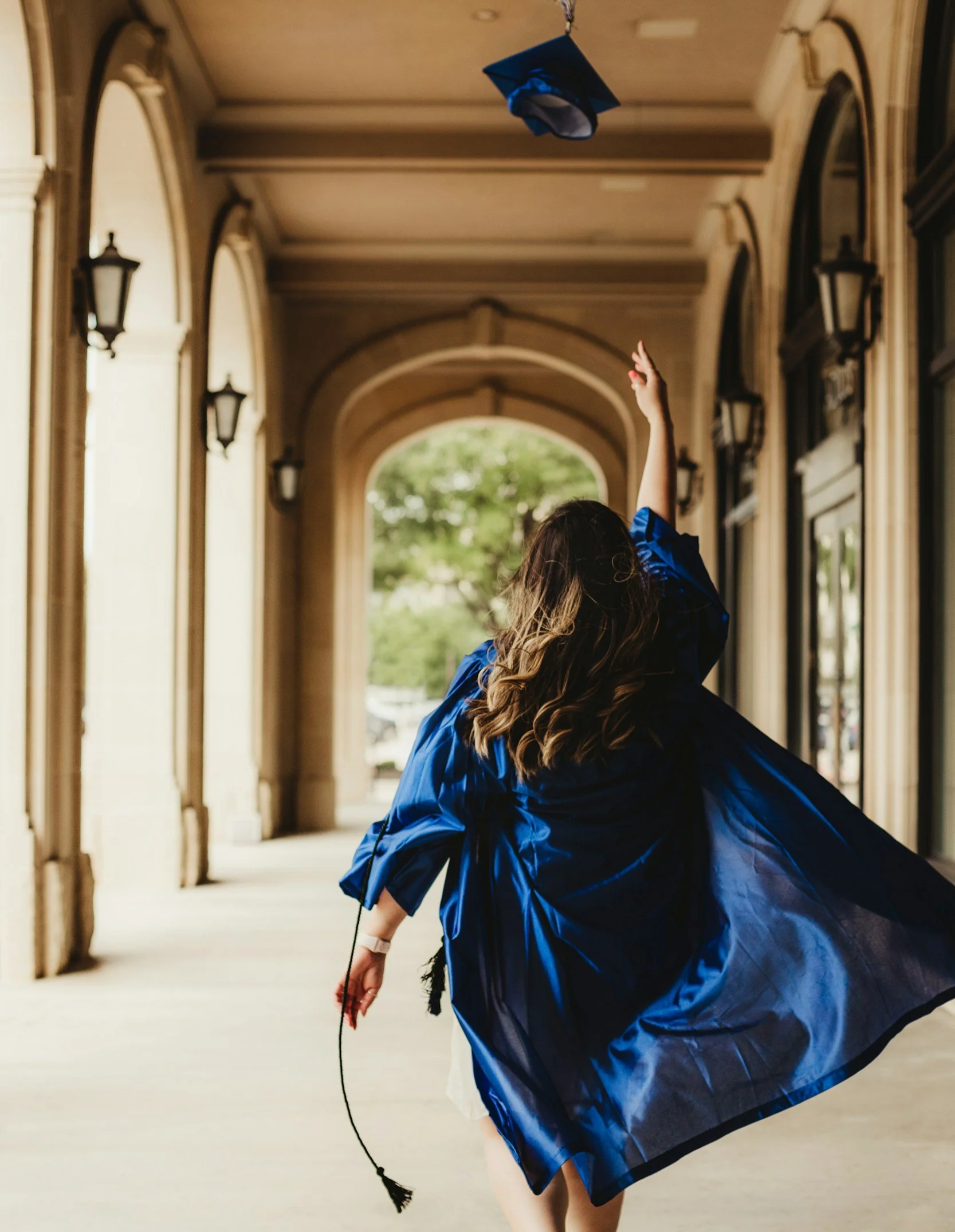 A graduate in a blue gown, seen from behind, runs down an arched stone walkway as her cap flies through the air above her.