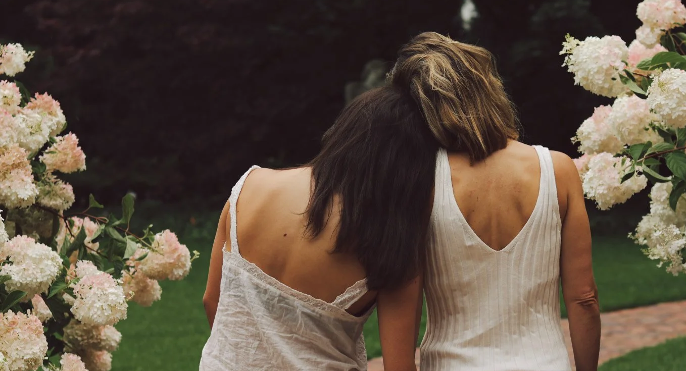 Seen from behind, a mother and daughter lean into each other on a brick path lined with pink and white hydrangeas.