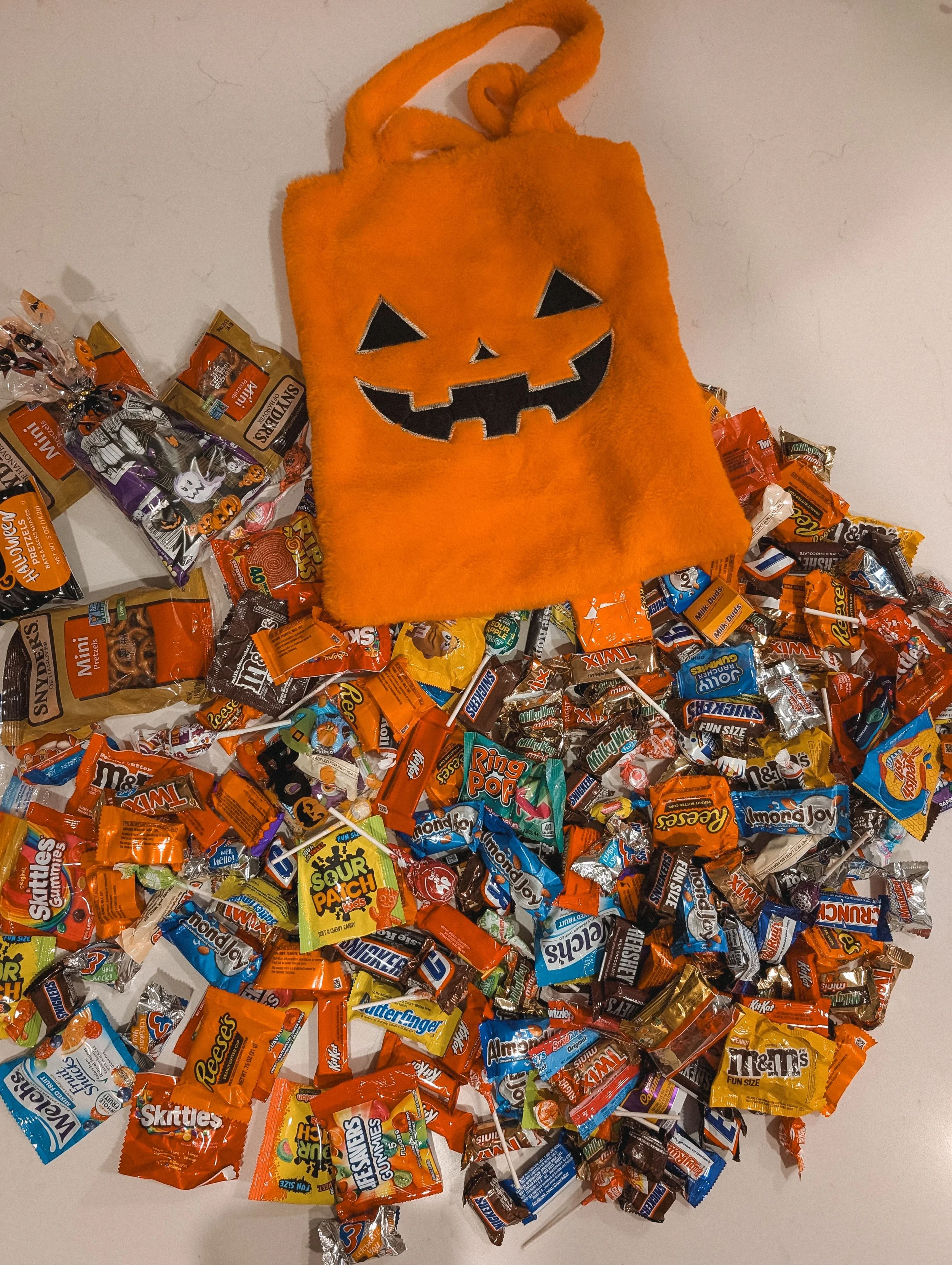 An orange jack-o'-lantern trick-or-treat bag surrounded by a huge pile of wrapped Halloween candy on a white surface.