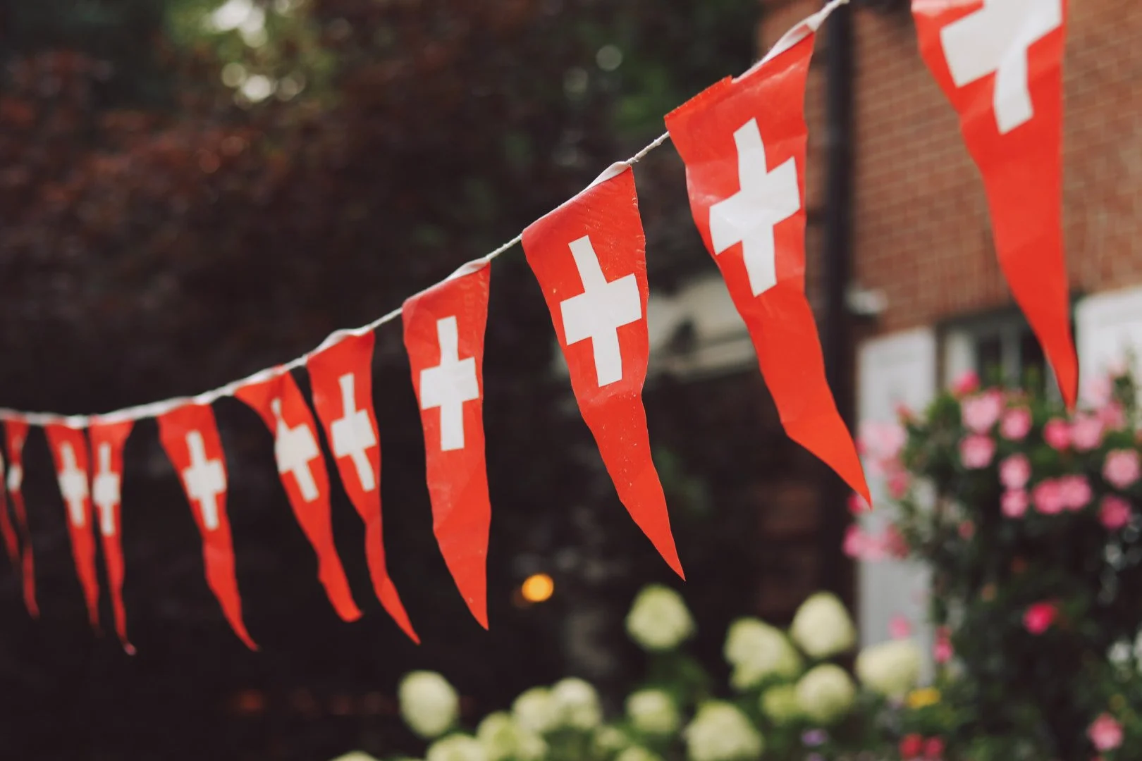 A string of red-and-white Swiss flag pennants hangs outdoors in front of a brick house and flowering garden.