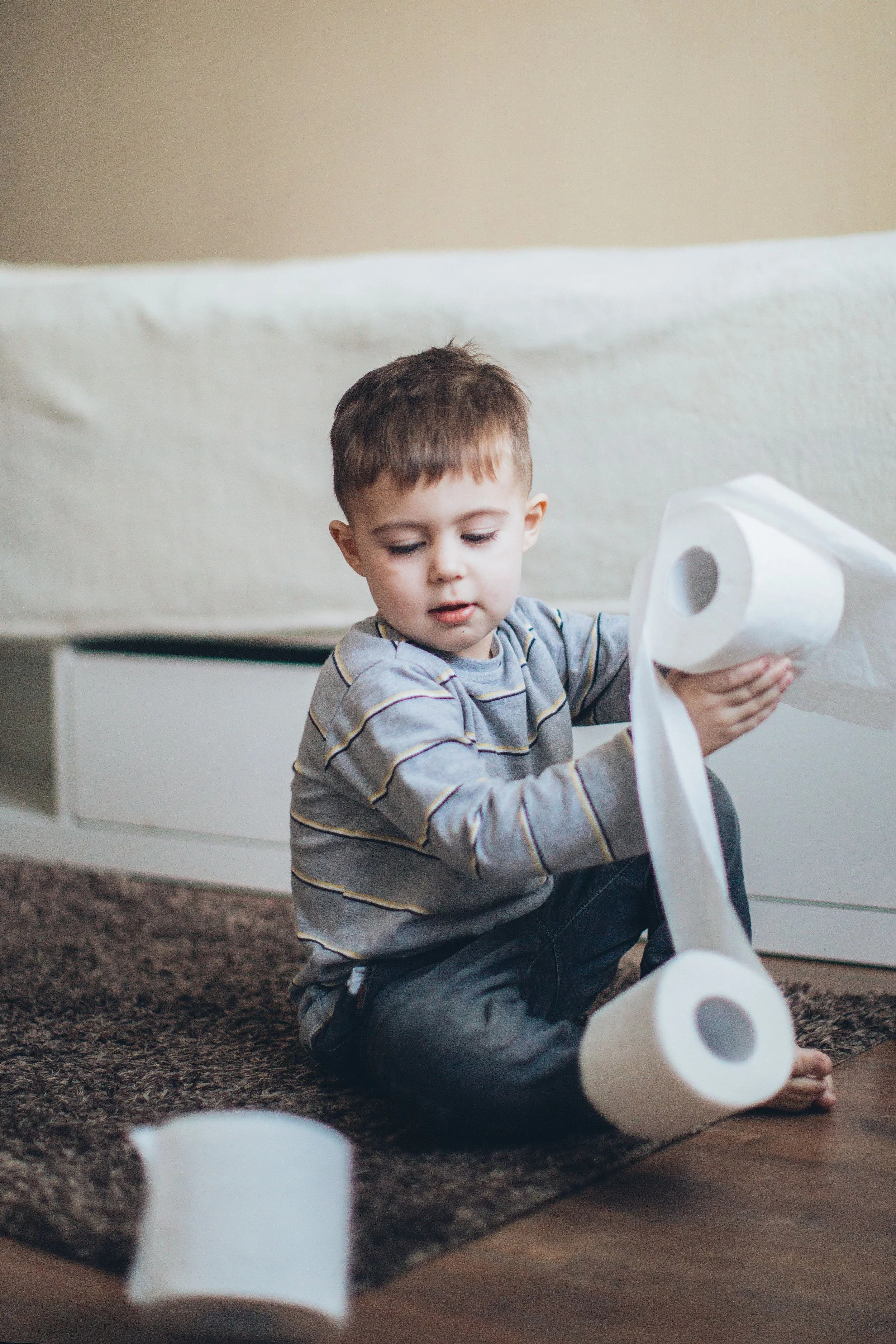 Young boy in grey striped shirt plays with two rolls of toilet paper on the ground.