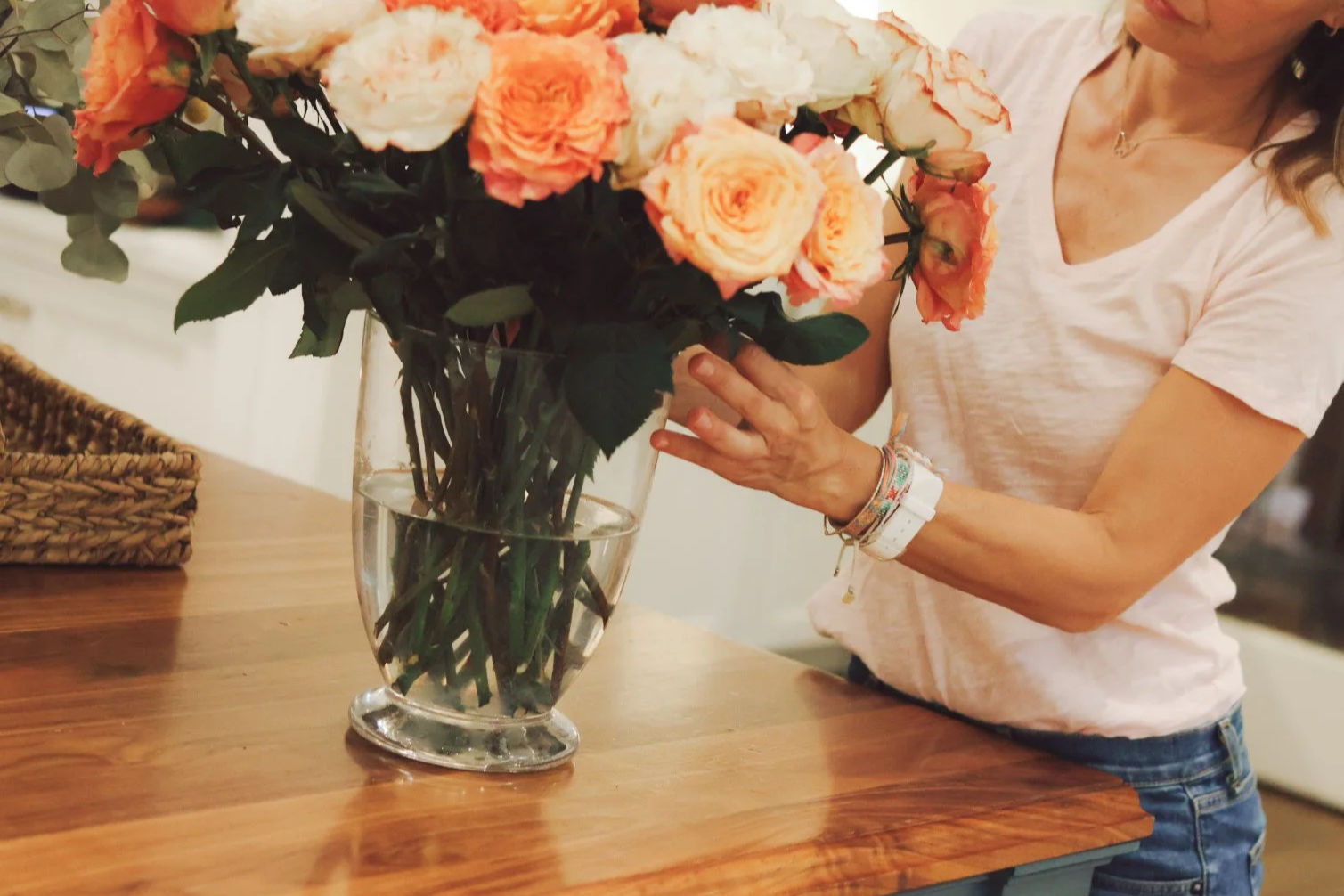 A mother in a light pink t-shirt and jeans arranges a bouquet of orange and cream roses in a glass vase on a wooden table.