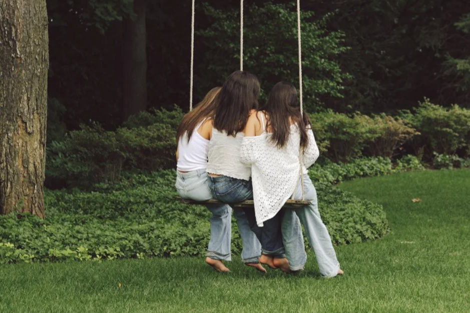 Seen from behind, three girls squeeze onto one tree swing, barefoot on the grass, shoulder to shoulder.
