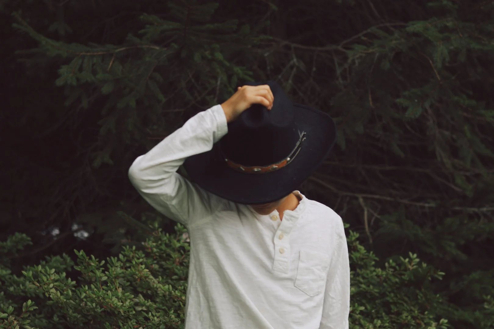 A young boy in a white shirt faces forward with his head tilted down, a wide black hat covering his face.