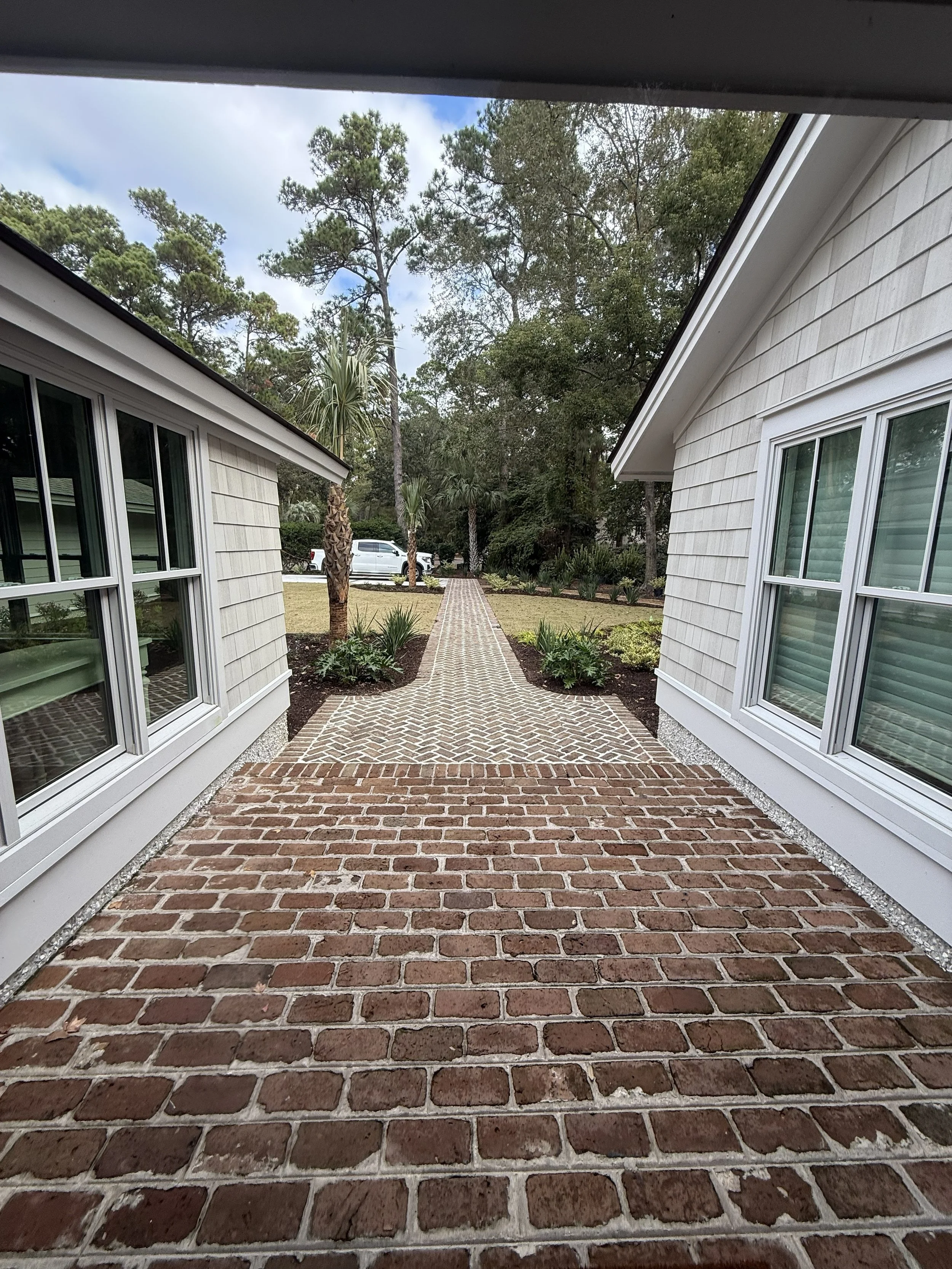 View of a brick pathway between two houses, with trees and a parked white truck in the background.
