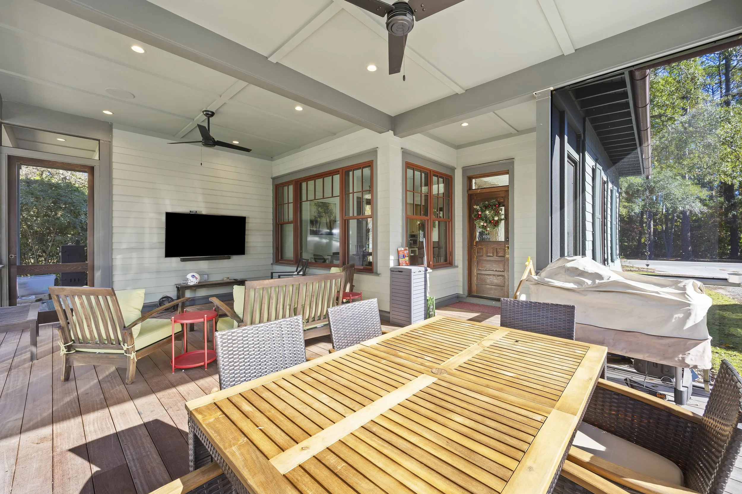 Covered patio area with wooden dining table, chairs, bench seating, a wall-mounted flat-screen TV, ceiling fans, string lighting, and outdoor equipment. The space overlooks a backyard with trees and a house decorated with a holiday wreath.