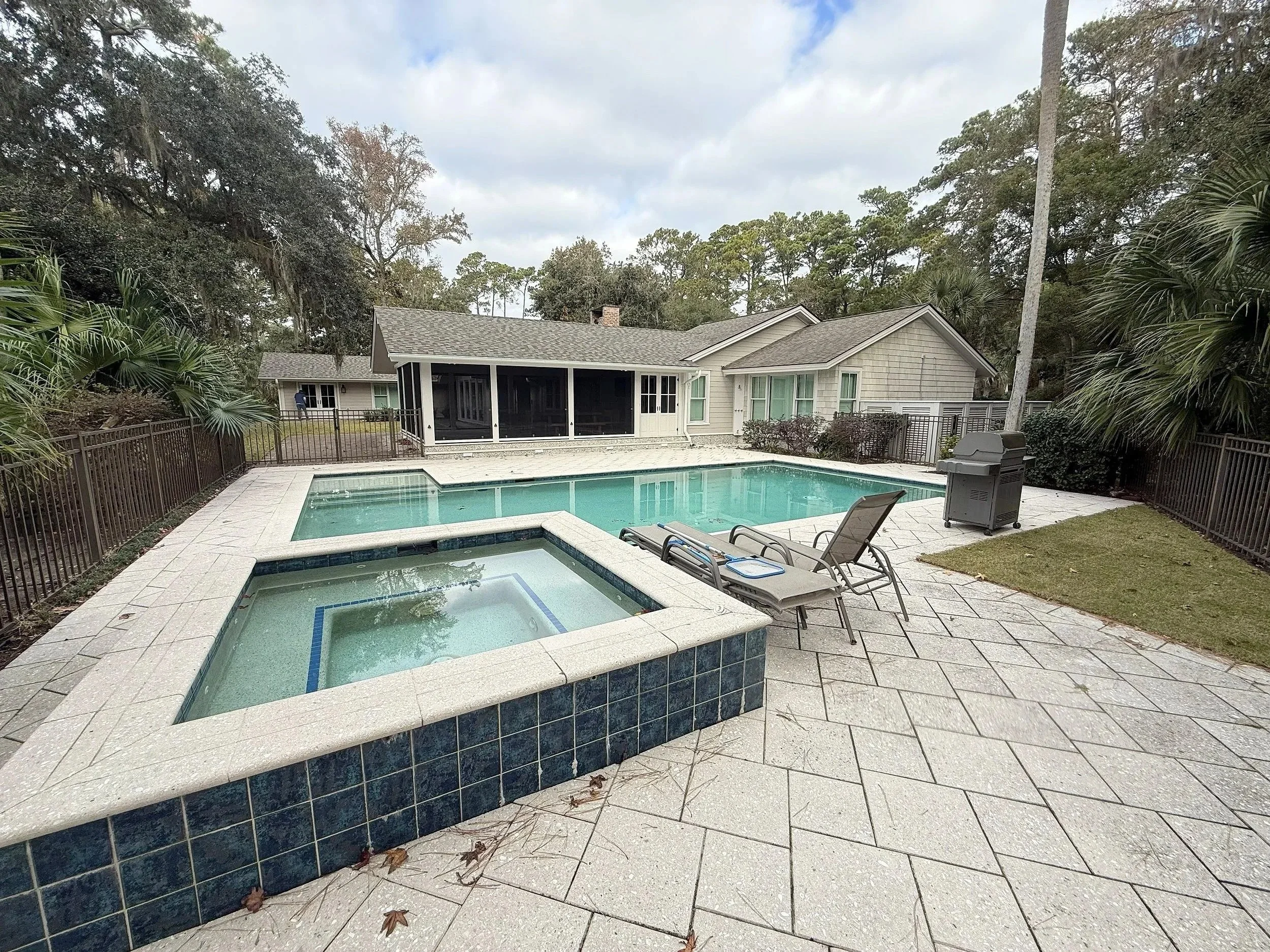 A backyard with a swimming pool, hot tub, and patio area enclosed by a black metal fence. There are two lounge chairs and a grill. A house with a screened porch, windows, and a roof is visible in the background, surrounded by trees and cloudy sky.
