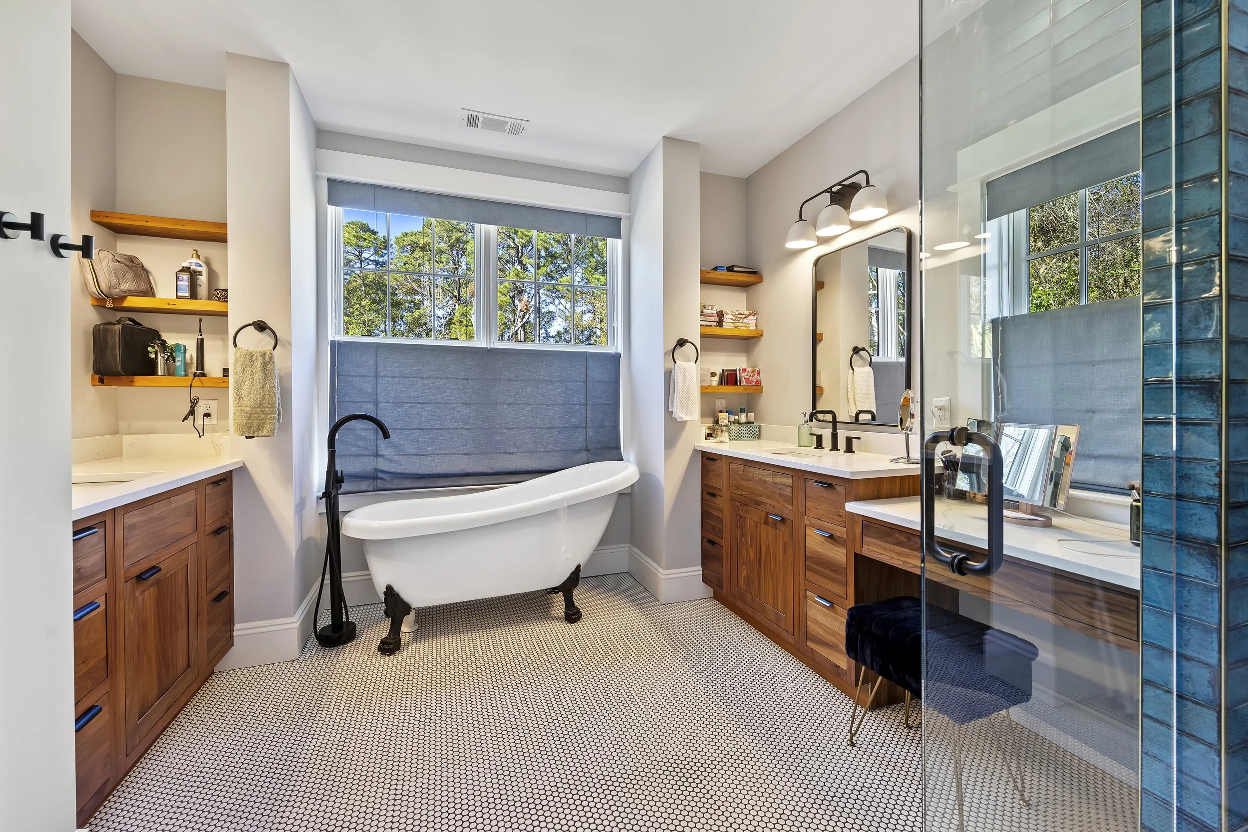 Modern bathroom with a clawfoot bathtub beneath a large window, wooden cabinetry, a vanity mirror with lights, open shelves with toiletries, and a walk-in shower with glass walls, all illuminated by natural light and wall-mounted fixtures.