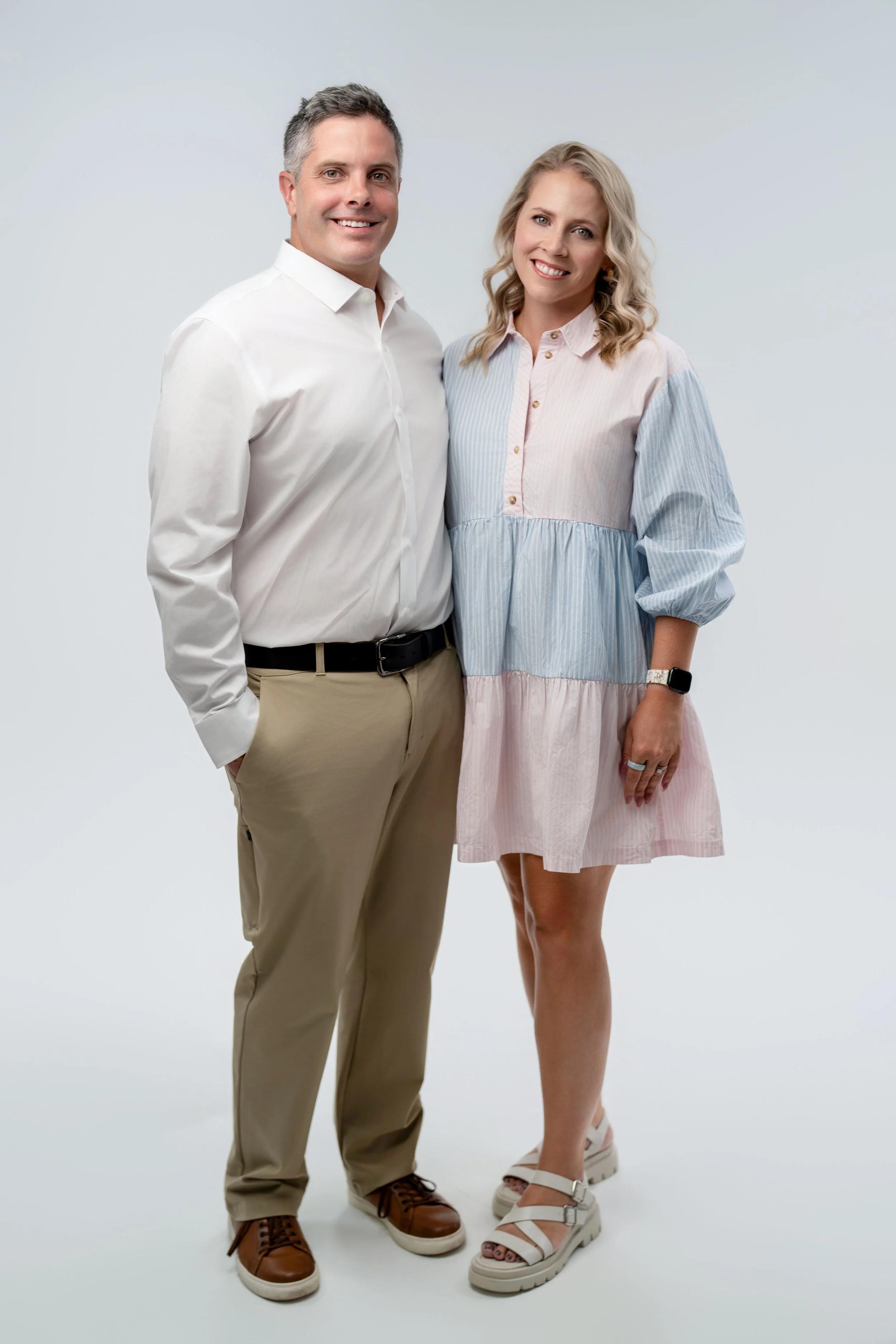 A smiling man and woman standing close together against a plain white background. The man, dressed in a white shirt and beige pants, has gray hair and brown shoes. The woman, wearing a pastel-colored patchwork dress and white sandals, has blonde wavy hair and a smartwatch on her left wrist.