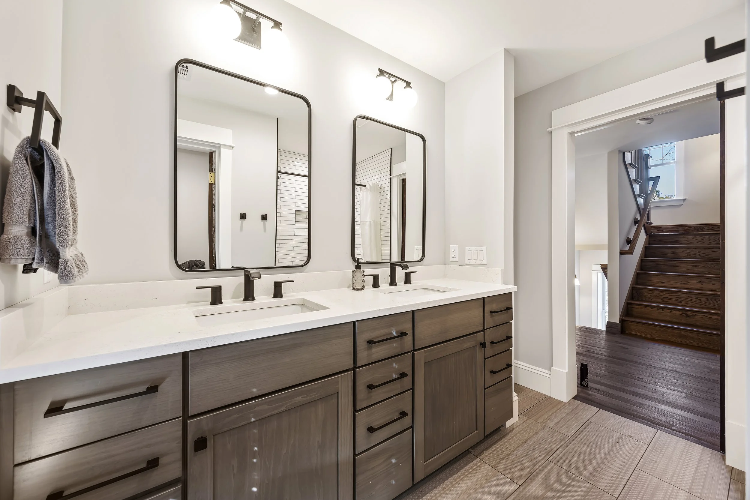 Modern bathroom vanity with two sinks, dark wood cabinets, large mirrors, and black fixtures, with a staircase and window visible in the background.
