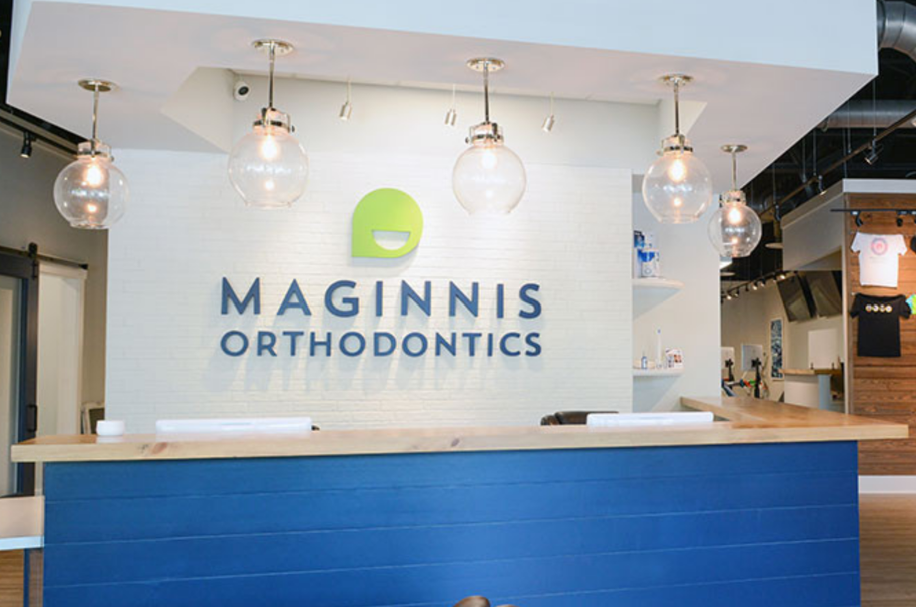 Interior view of an orthodontic clinic with a white brick wall featuring the blue and green logo of Maginnis Orthodontics. Five glass globe pendant lights hang from the ceiling. Modern decor and a reception area with a wooden counter are visible.