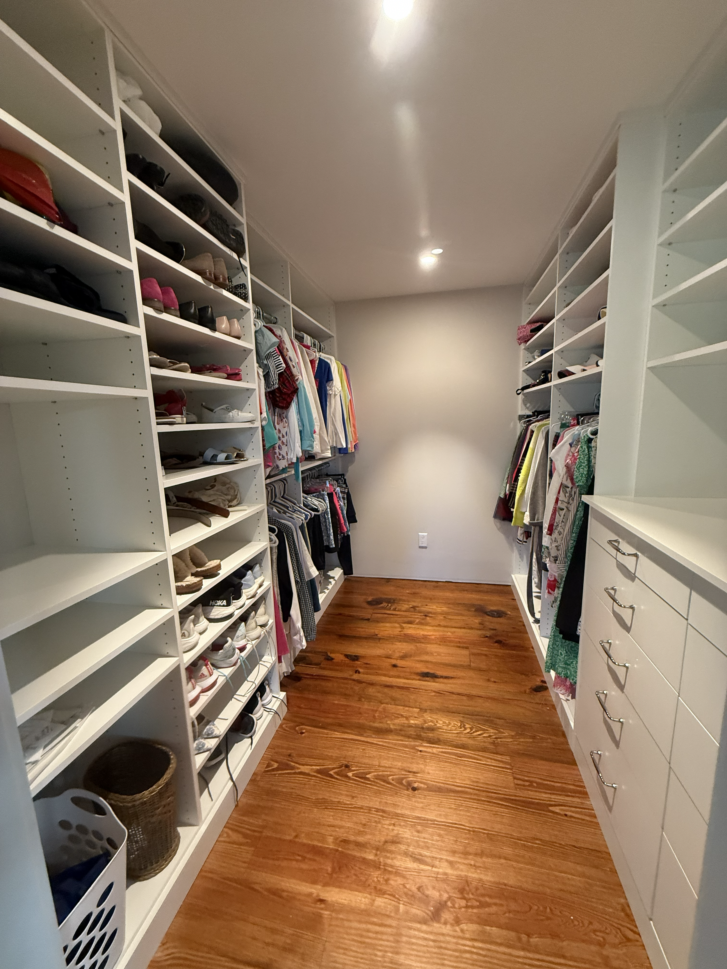 A walk-in closet with white shelving and hanging clothes, shoes, and accessories organized on the shelves and racks, with hardwood flooring.