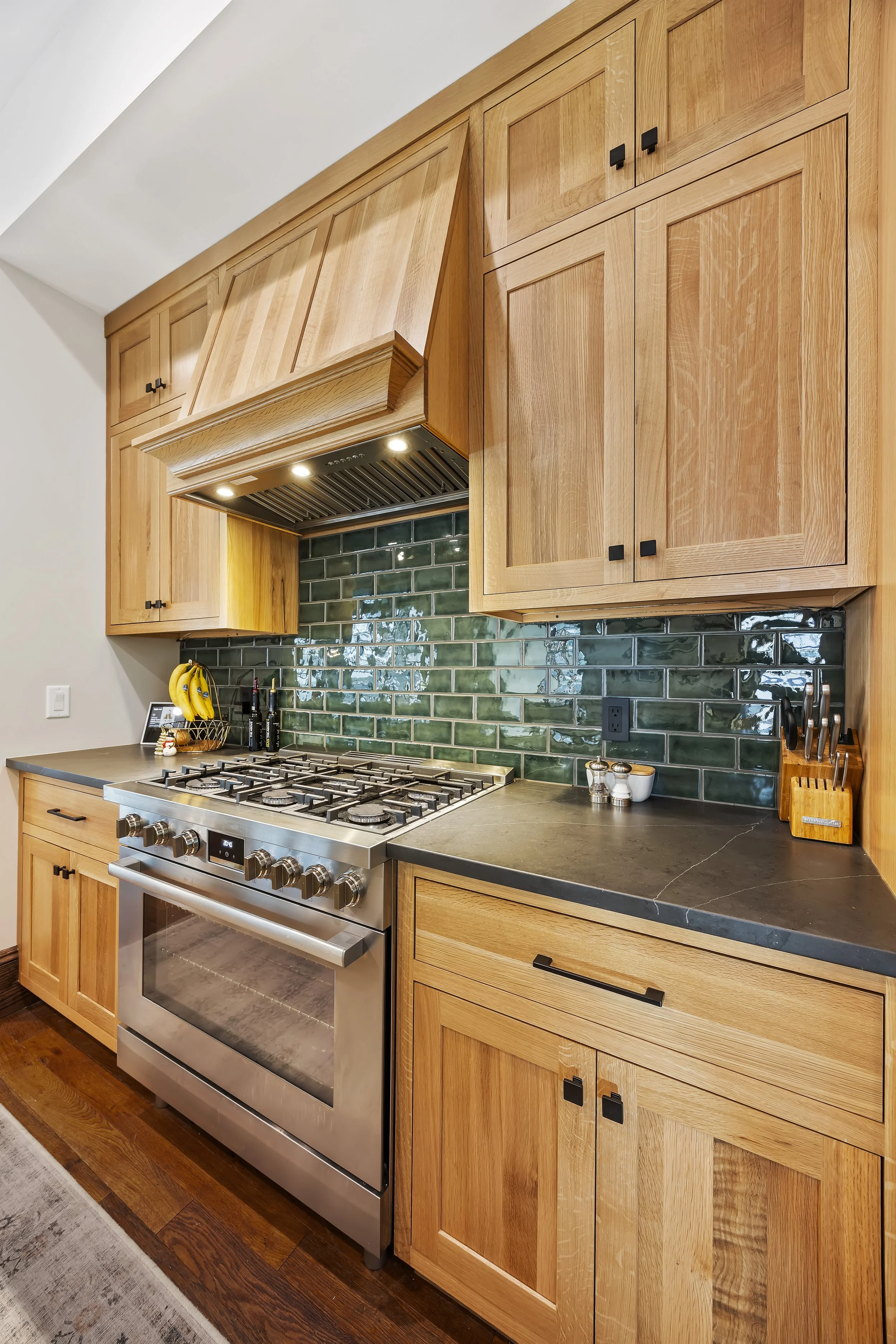 Kitchen with wooden cabinets, a green tile backsplash, and a stainless steel stove. Small appliances and utensils are on the counter, including a bunch of bananas.