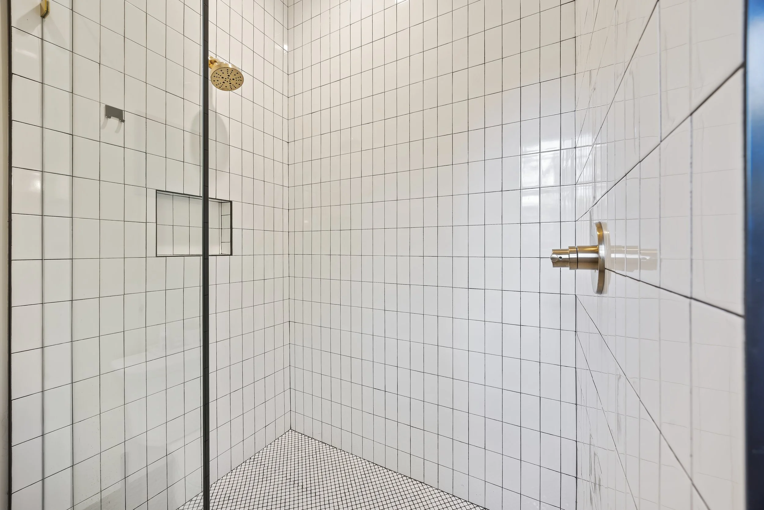 A modern shower with white square tiles on the walls and small black and white mosaic tiles on the floor. There is a round, gold-colored showerhead mounted on the ceiling and a small rectangular built-in shelf in the wall. A glass panel separates the