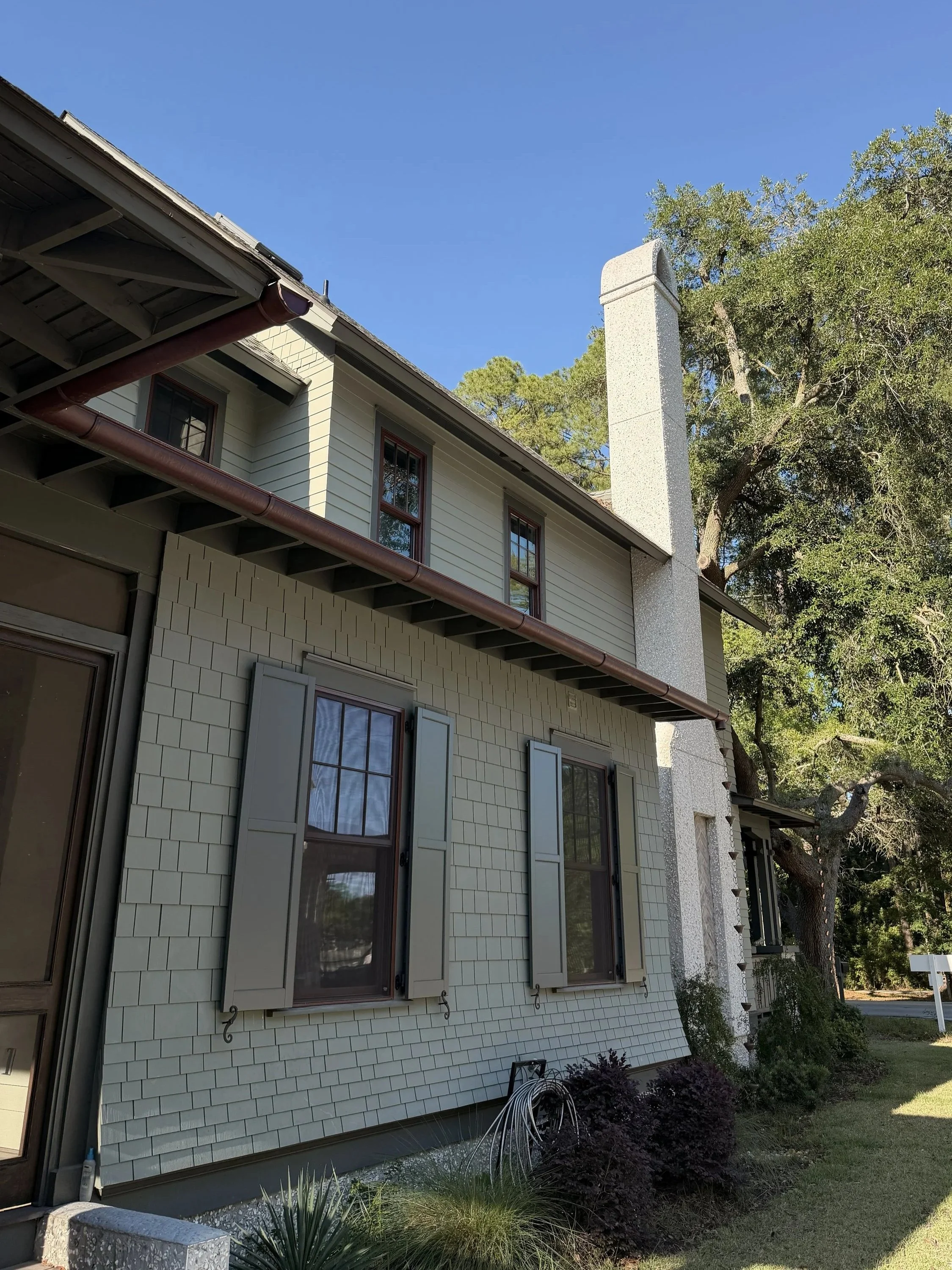 The image shows the exterior of a two-story house with cream-colored siding and brown window frames. There are multiple windows with shutters, a gutter system, and a chimney extending above the roofline. The yard features bushes and grass, with tall trees in the background and a clear blue sky.