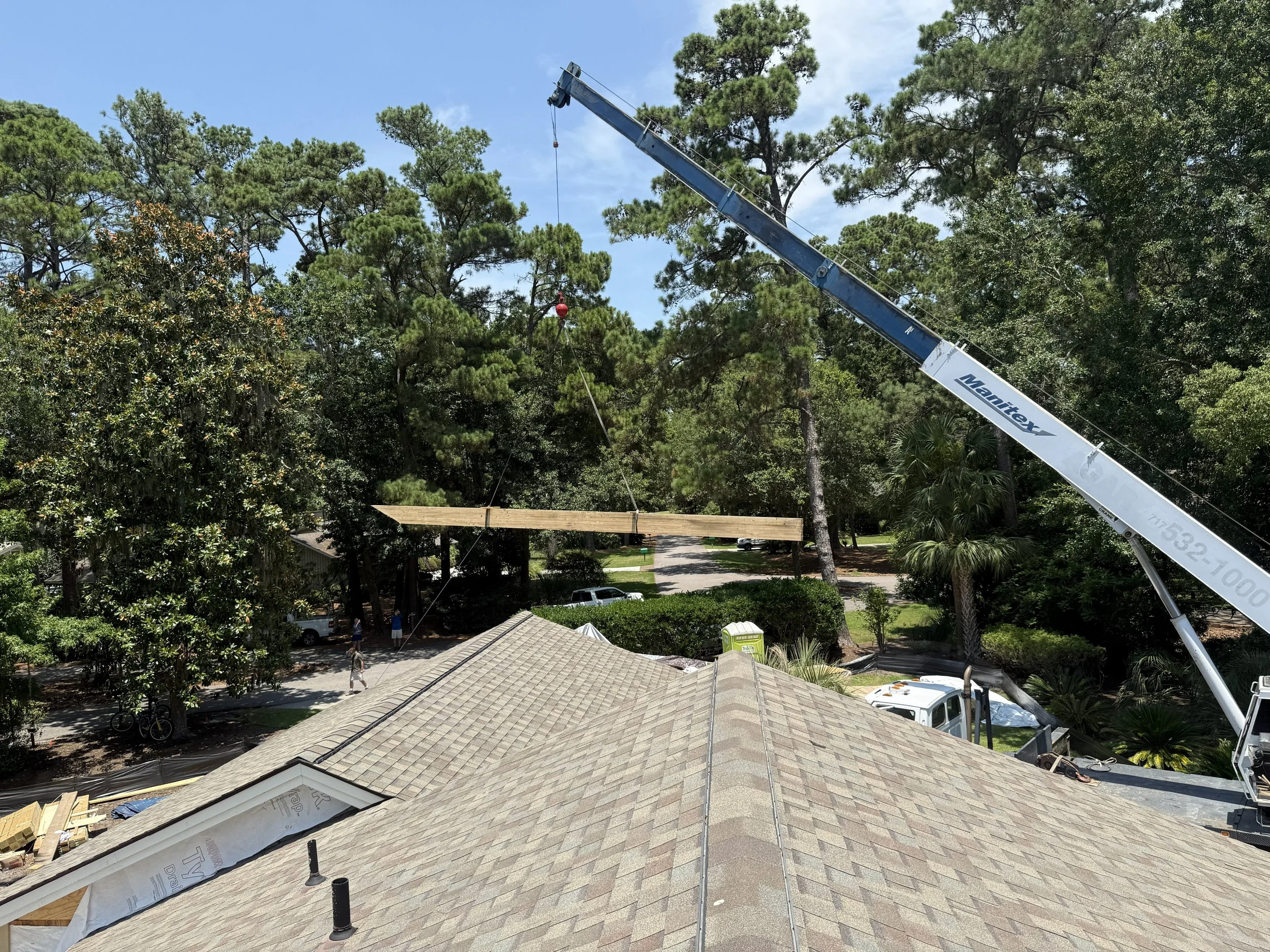 View from a house roof under construction, with a crane lifting a large wooden beam, surrounded by tall trees and a blue sky.