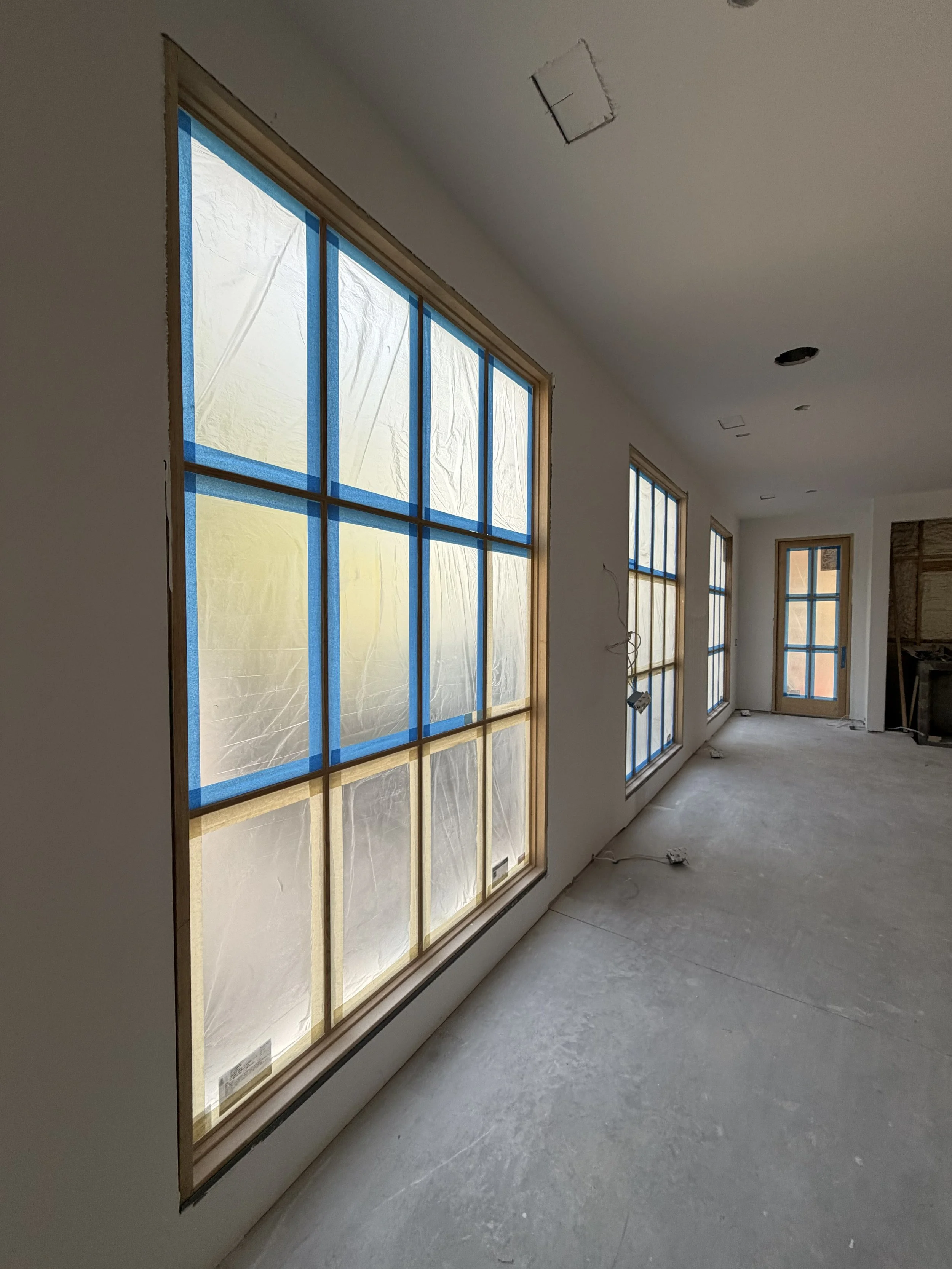 Living room under construction with large windows covered in protective plastic and blue painter's tape, unfinished walls and ceiling, and electrical wiring visible.