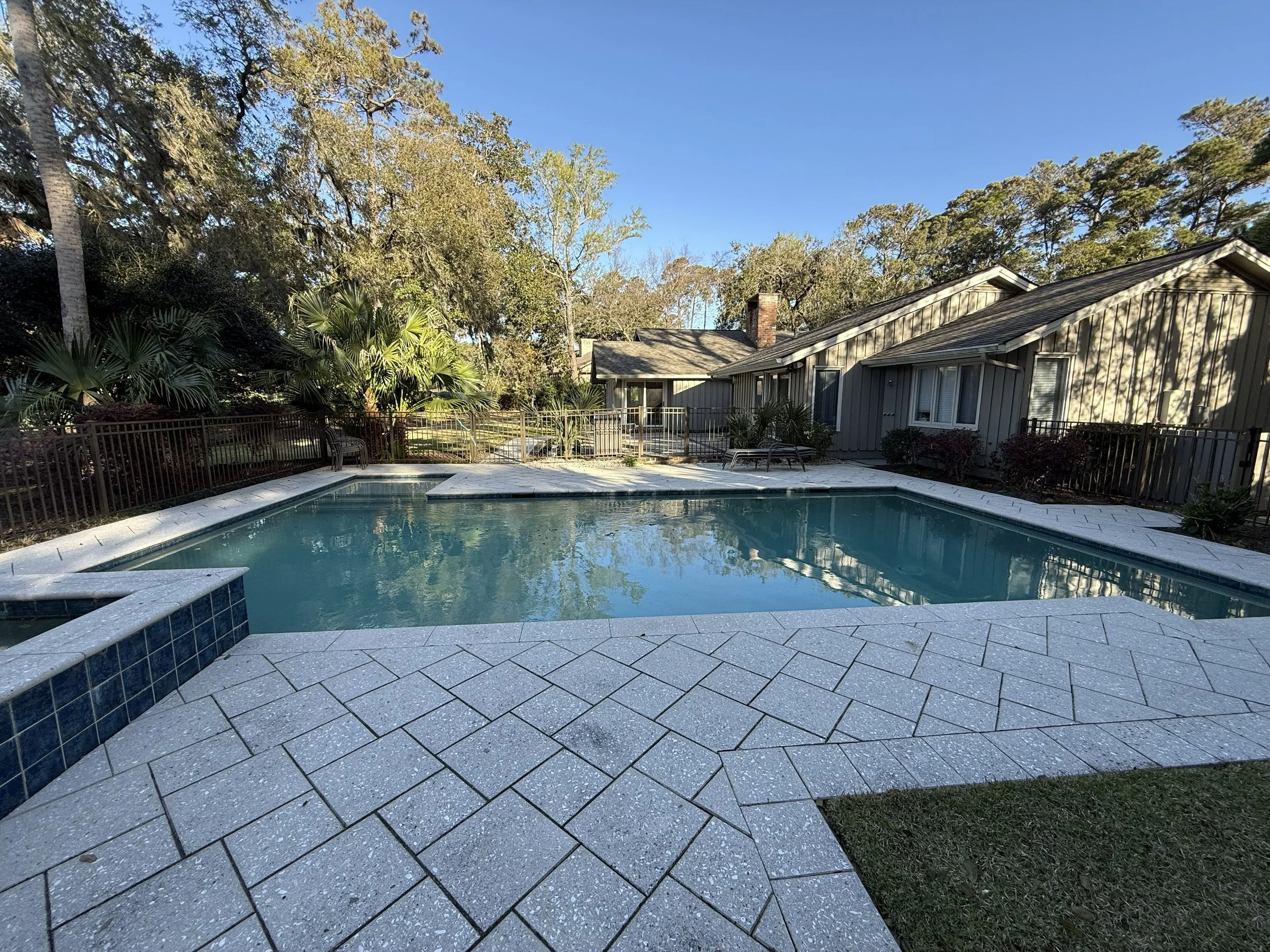 Residential backyard with a swimming pool, surrounded by a tiled patio, various plants, and trees, and a house with a brick chimney in the background.