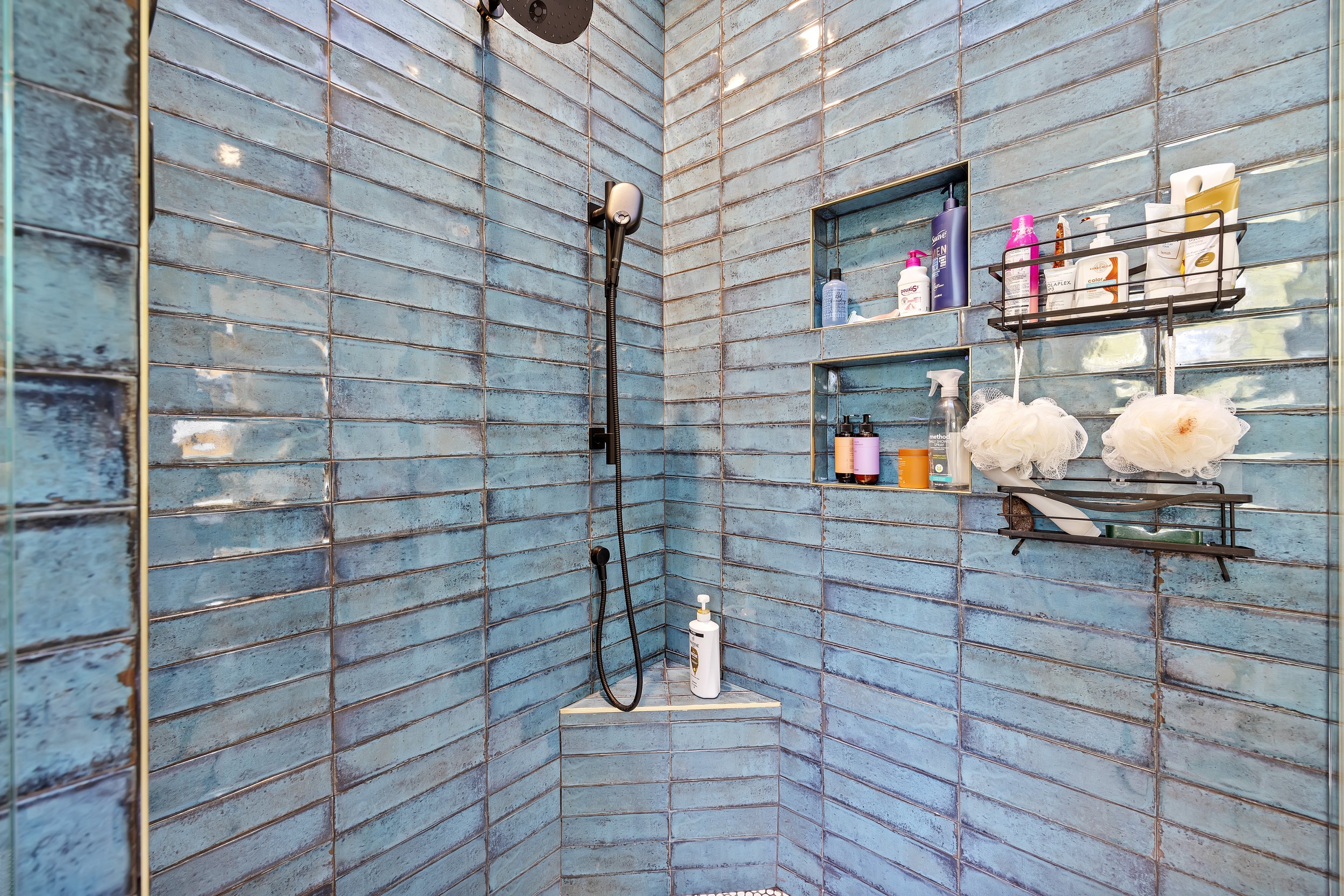 A corner shower with blue distressed tiles, a black wall-mounted showerhead, and three built-in shelves holding toiletries and bath products.