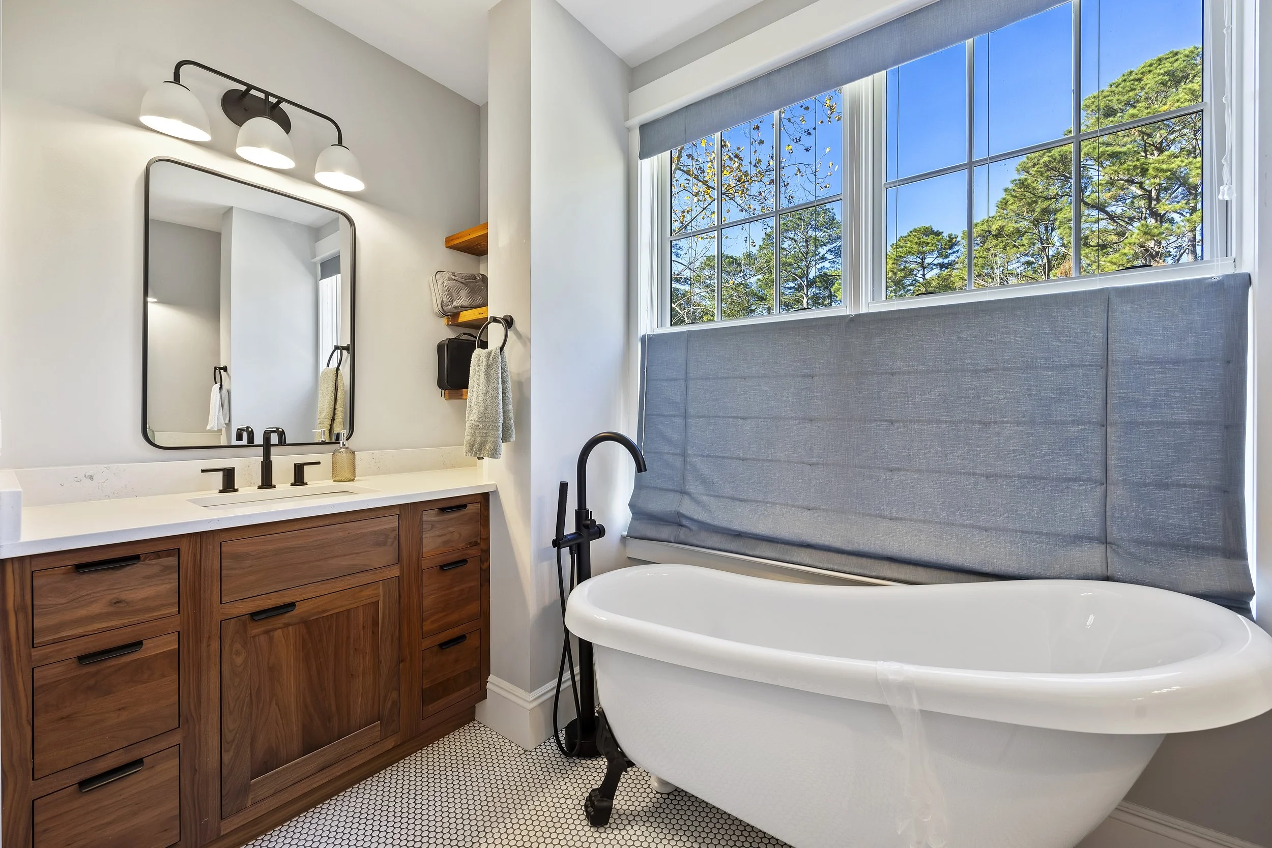 A bathroom with a wooden vanity, black faucet, large mirror, and a bathtub with a gray cushioned headrest. There are three white globed light fixtures above the mirror, two wooden shelves with a toiletries bag and towel, and a window with blue blinds