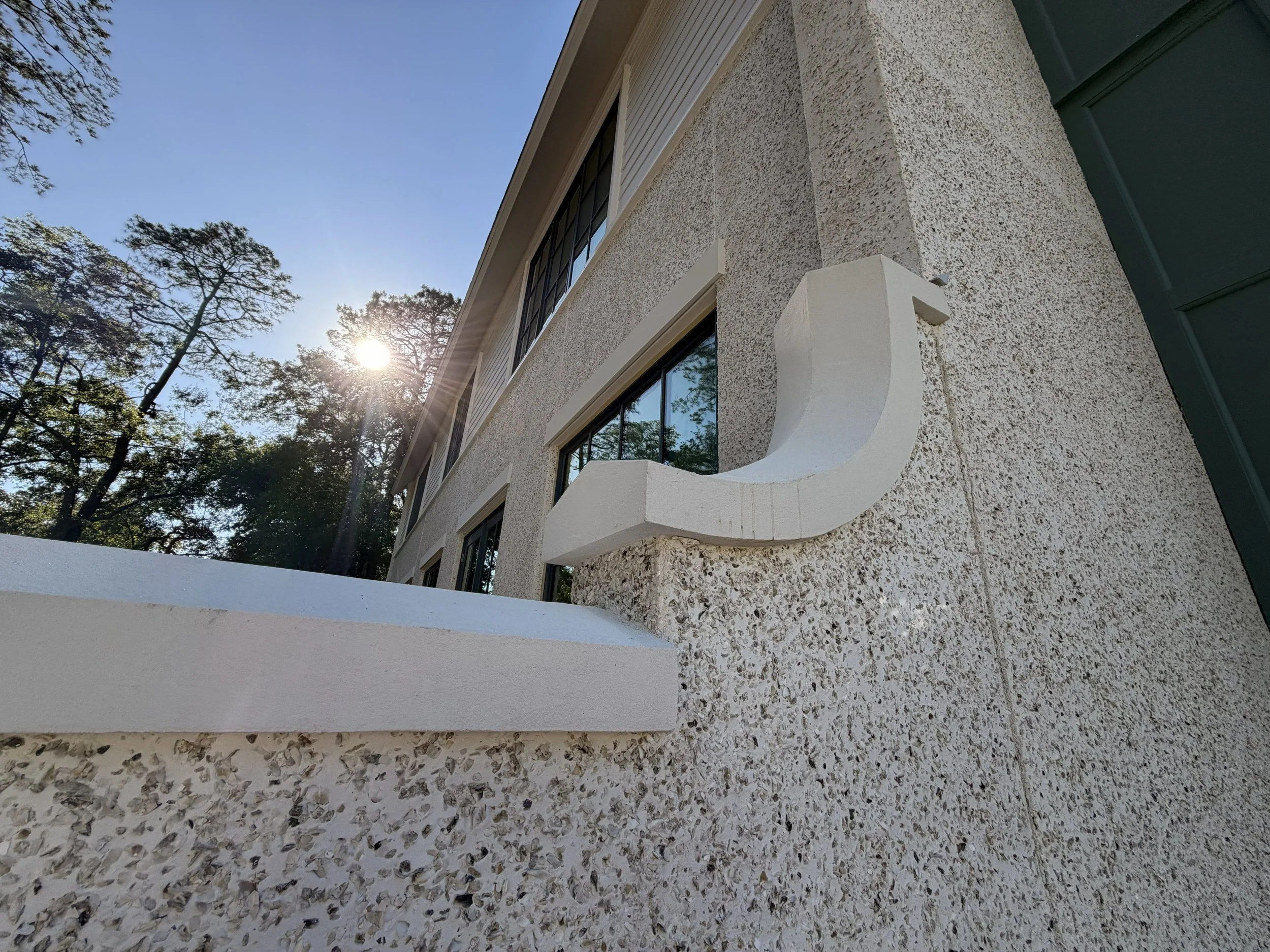 Close-up of a building's exterior wall with textured stucco finish, windows, and a white curved decorative feature under the window. Trees and the sun are visible in the sky.