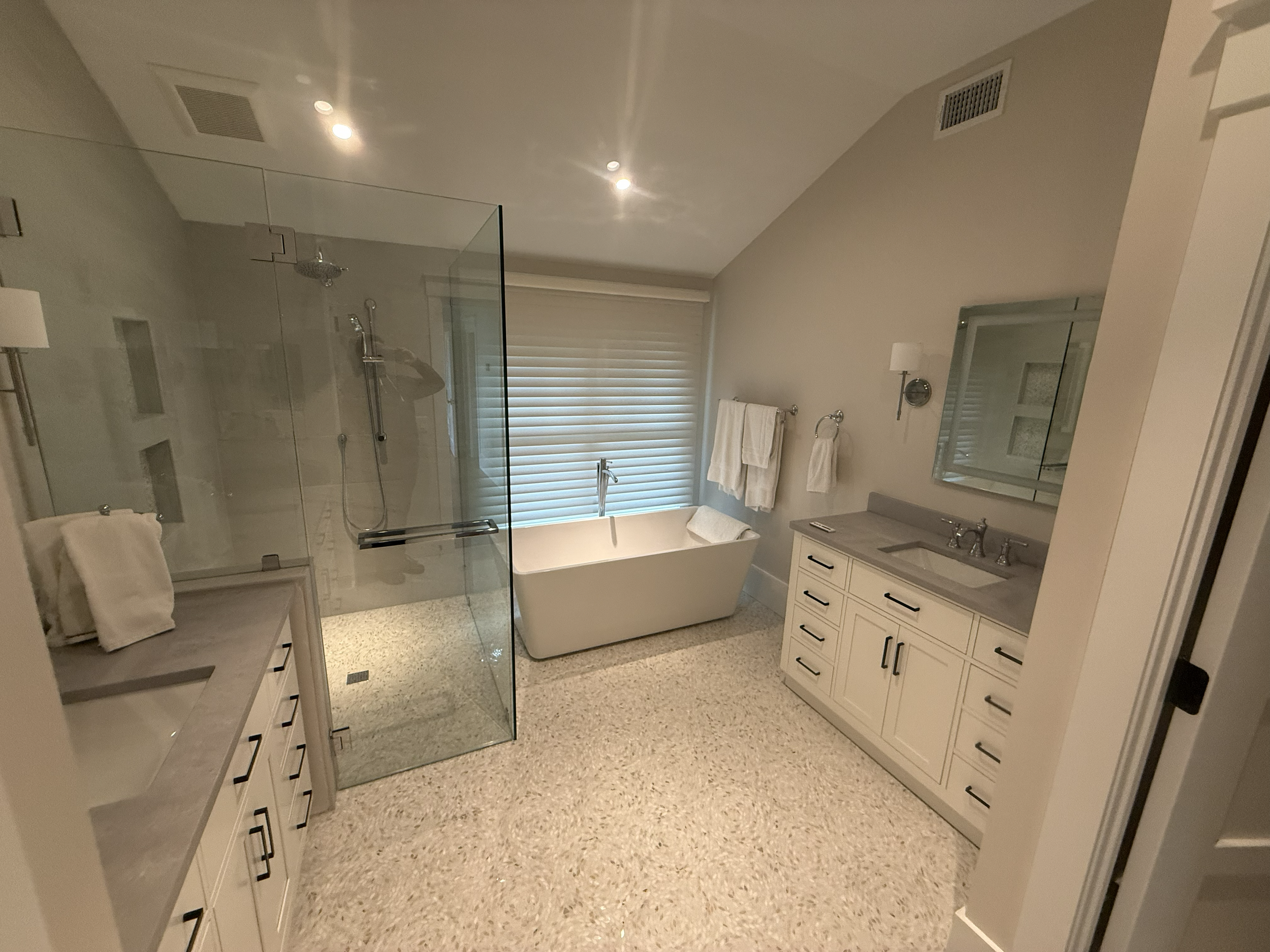 A modern bathroom with a glass shower enclosure, a free-standing bathtub, white cabinetry, a mirror, and a window with blinds.