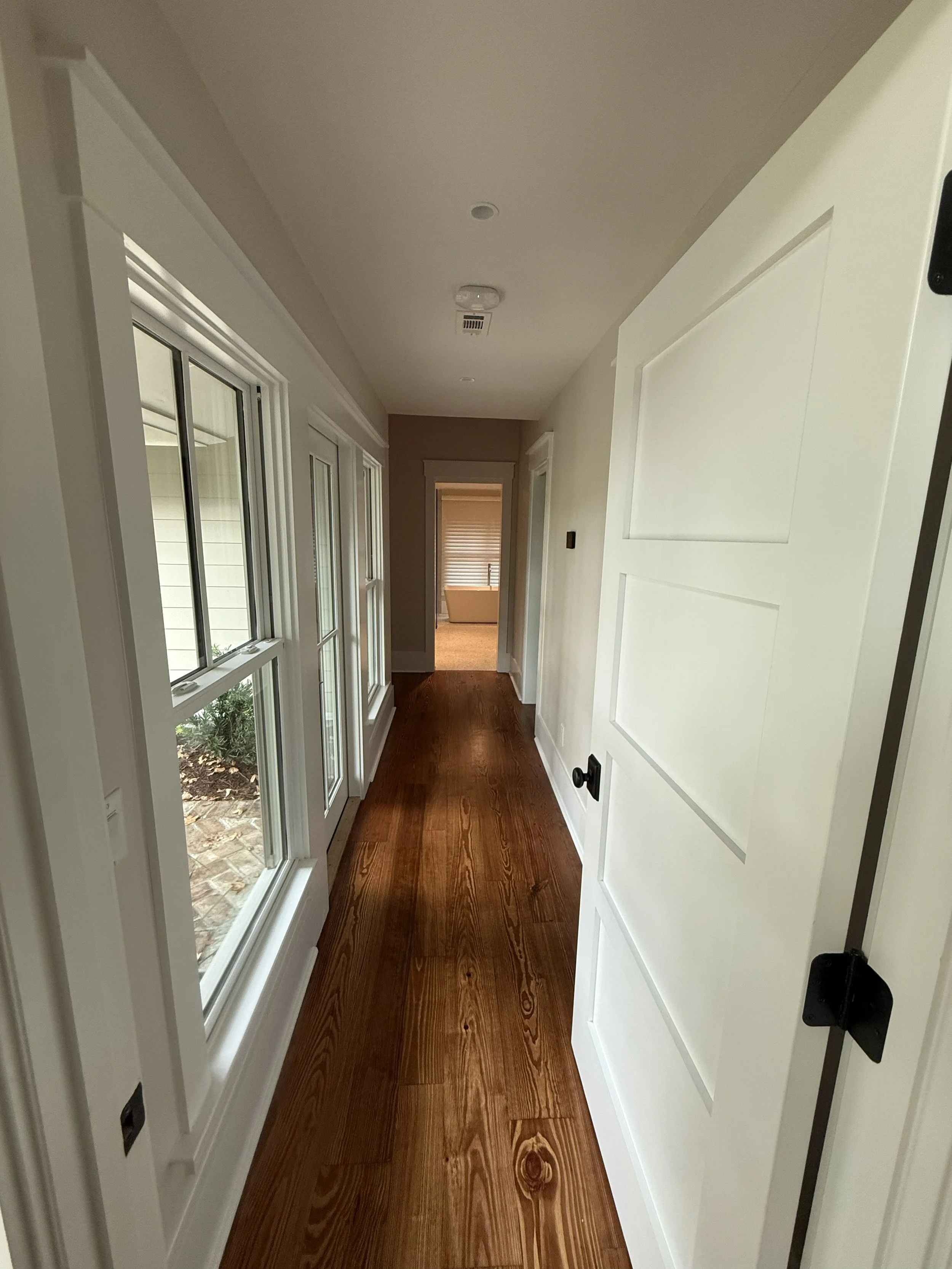 A hallway with a floor made of polished wood, white walls, a large window on the left showing an outdoor space, and a partially open door on the right. In the distance, a room with a bathtub is visible.