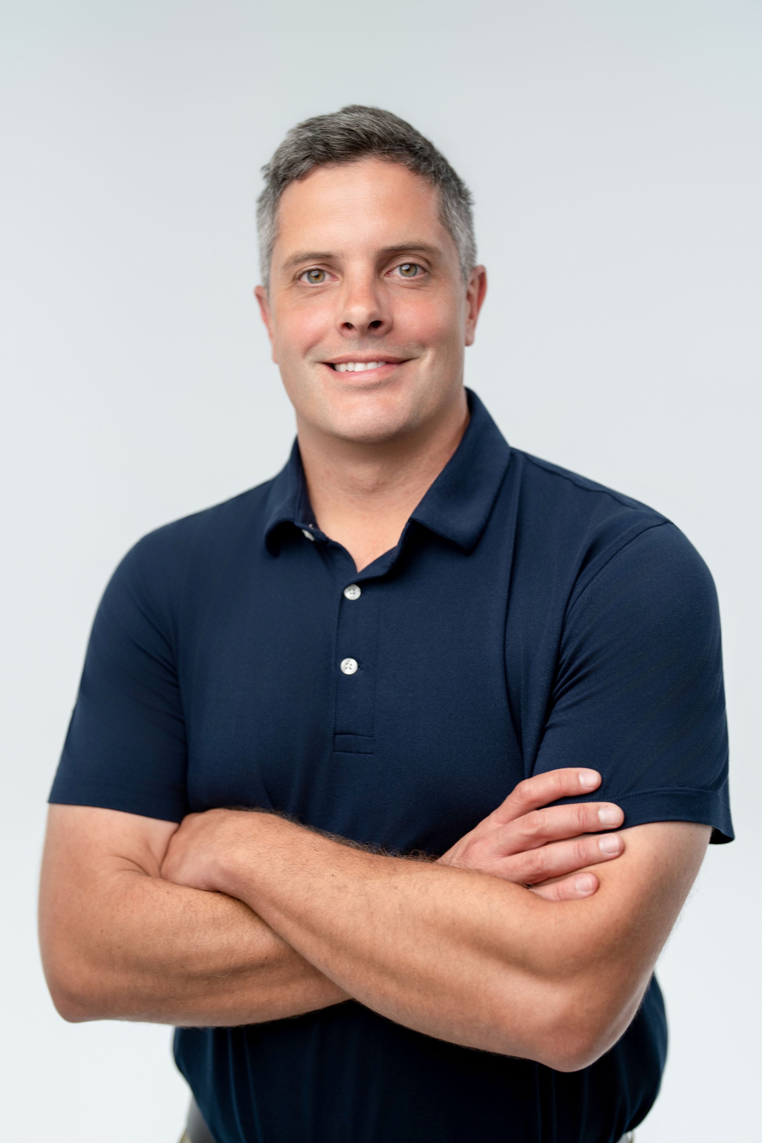 A man with short gray hair and green eyes, smiling, wearing a navy blue polo shirt with arms crossed in front of a plain light gray background.