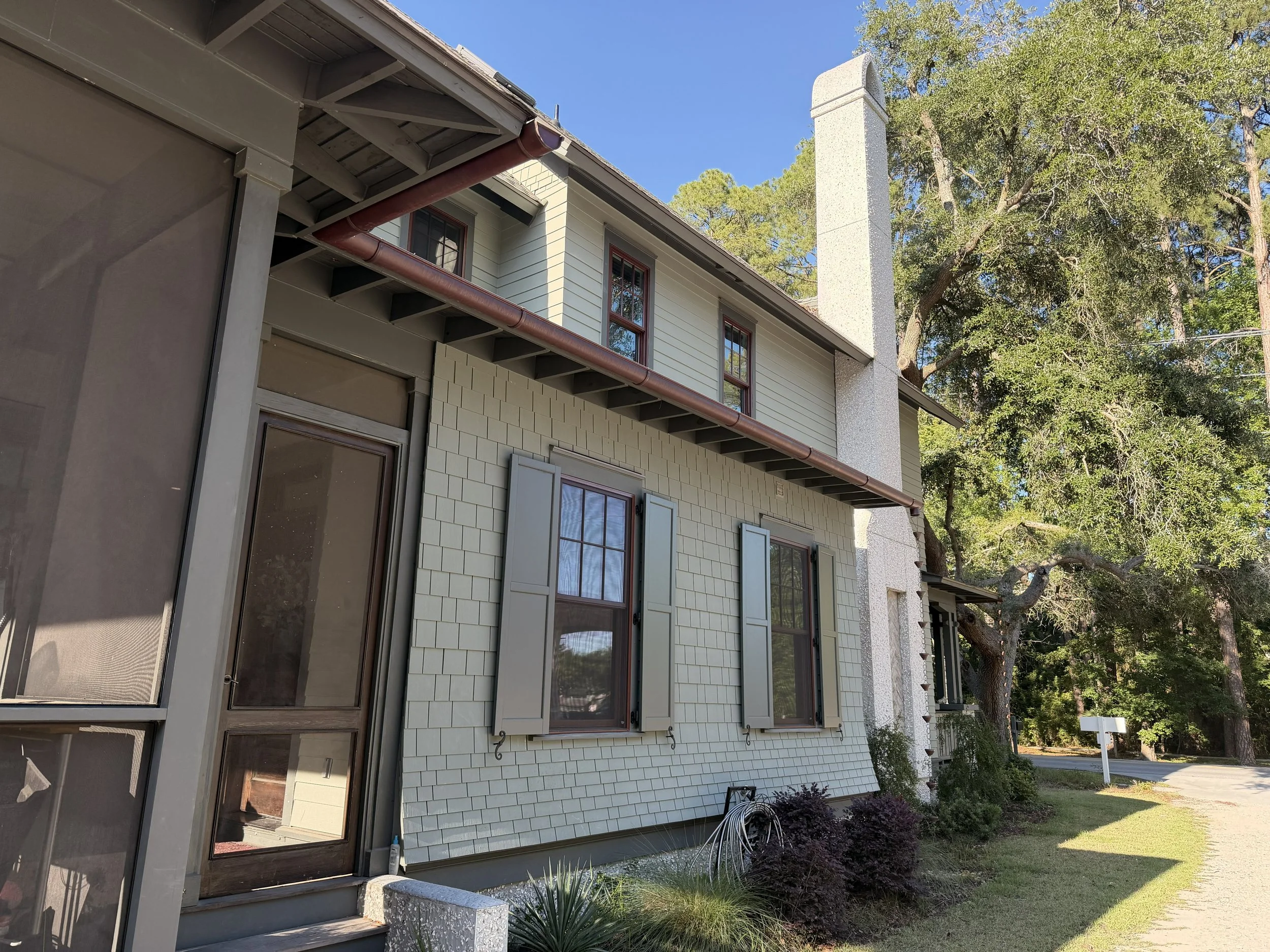 View of a house's exterior with white siding, large windows with shutters, a brick foundation, and a backyard with grass, trees, and plants, under a clear blue sky.