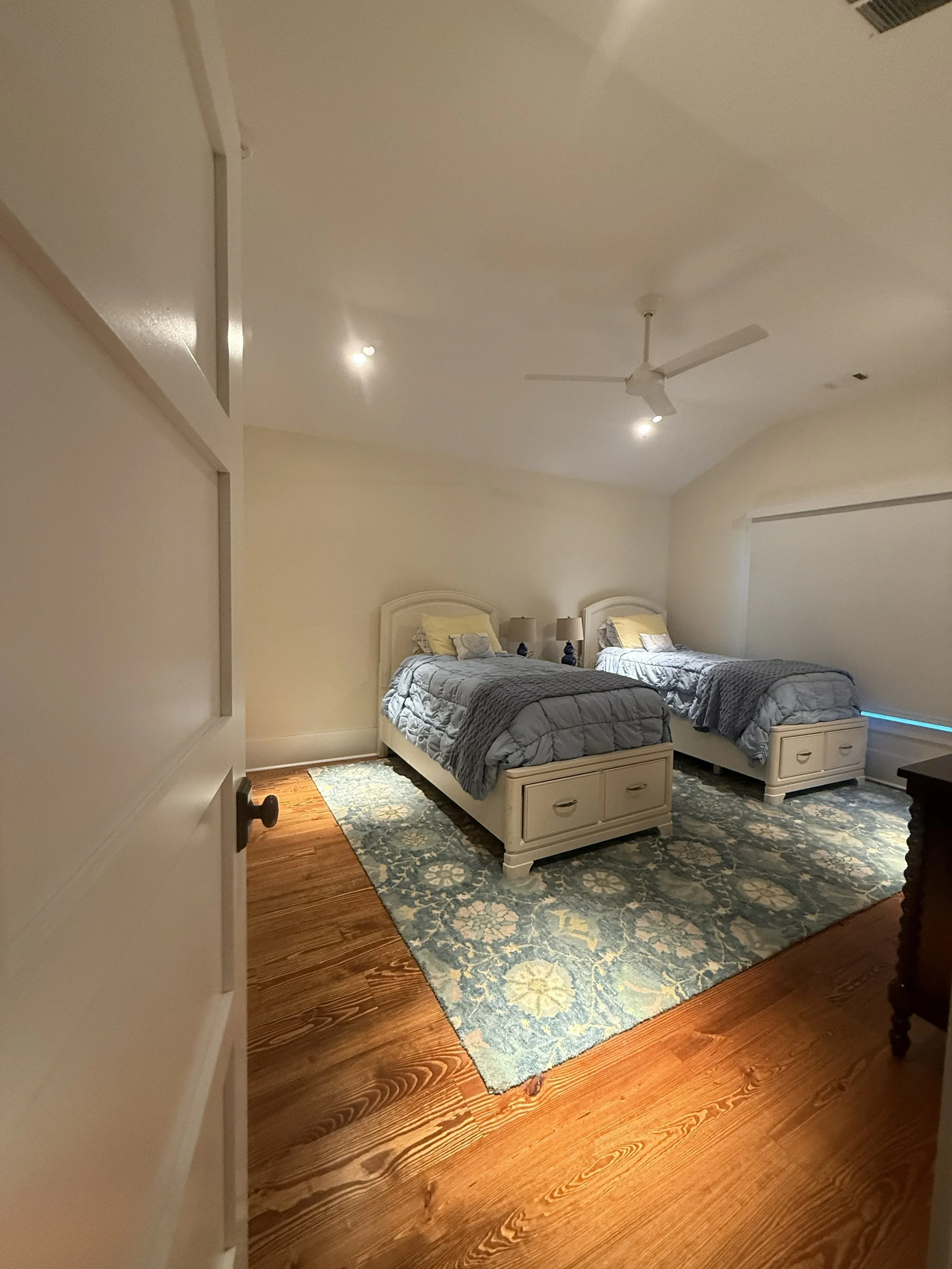 A bedroom with two twin beds, blue and beige bedding, a rug on hardwood floor, and a window with blinds.