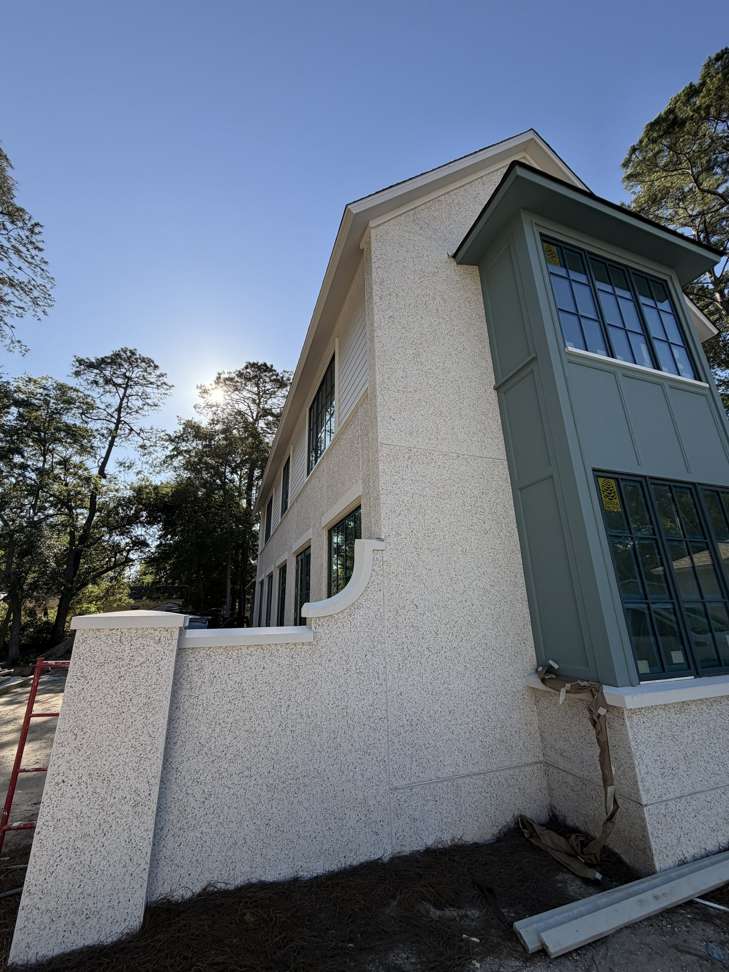 Side view of a modern house under construction with textured white exterior walls, large windows with black grids, and a green bay window. The sun is shining in the background, and there are tall trees around the house. Construction materials are on 