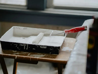 Paint tray with white paint, red-handled paint roller, paintbrush on a wooden table with paint splatters, near a window.