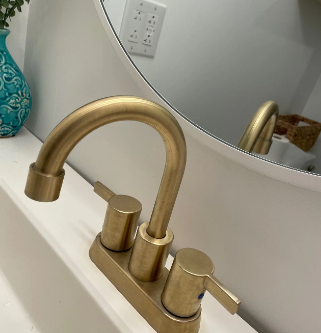 A bathroom sink faucet with a brushed gold finish, mounted on a white countertop, with a round mirror above and a decorative turquoise vase in the background.