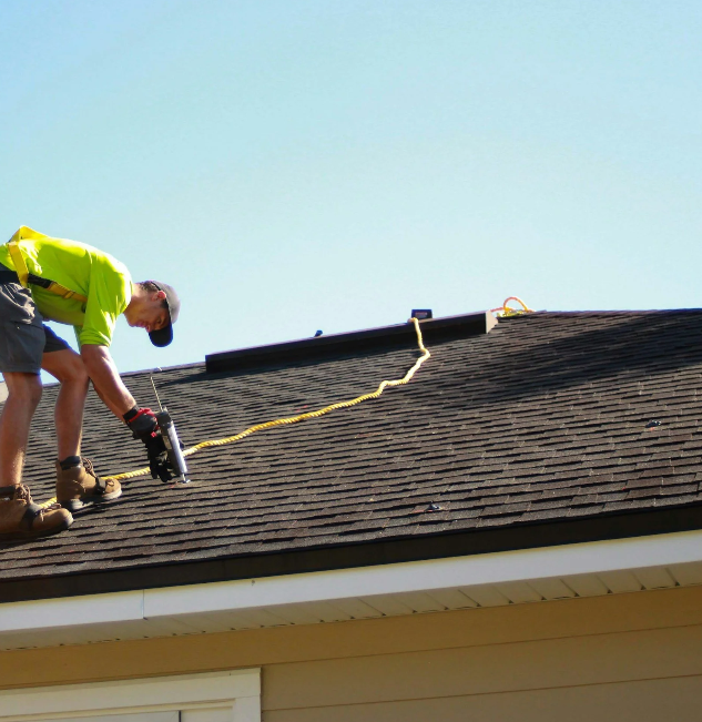 A worker on a roof using a power drill to secure shingles.