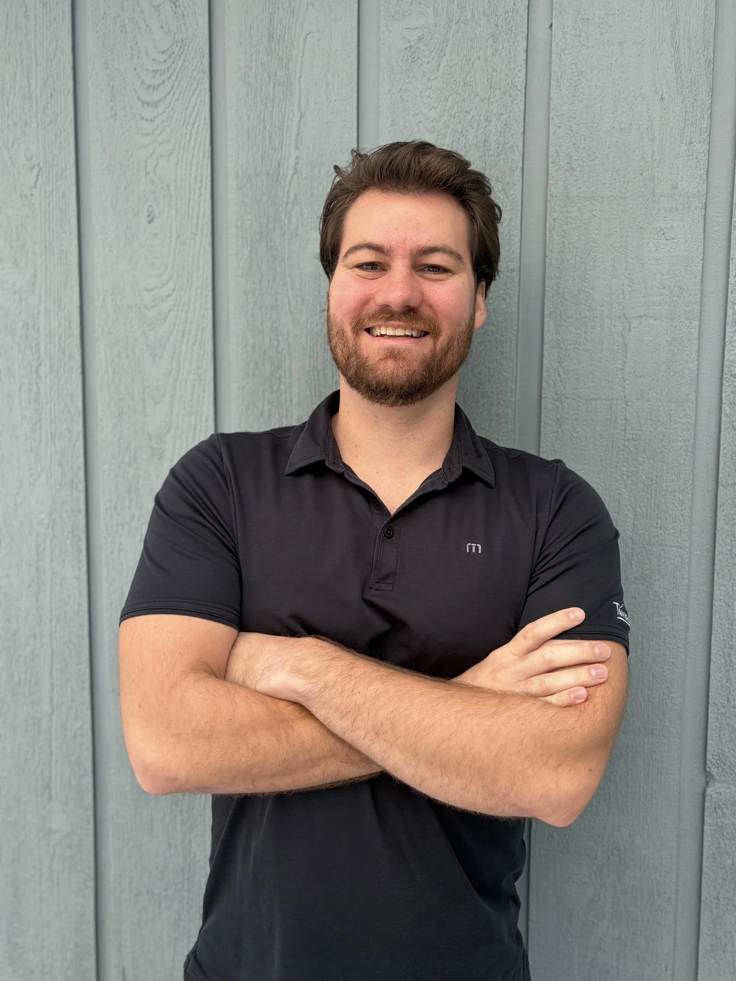 A smiling man with dark hair and a beard, wearing a black polo shirt and standing with arms crossed in front of a light green wooden wall.