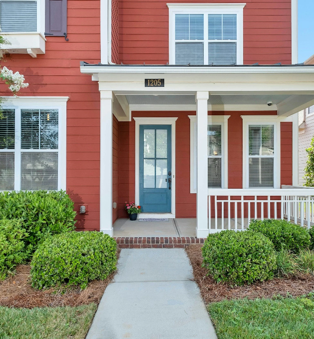 Front porch of a red house with a blue front door, white trim, and a small flower pot on the porch, surrounded by green bushes and shrubs.