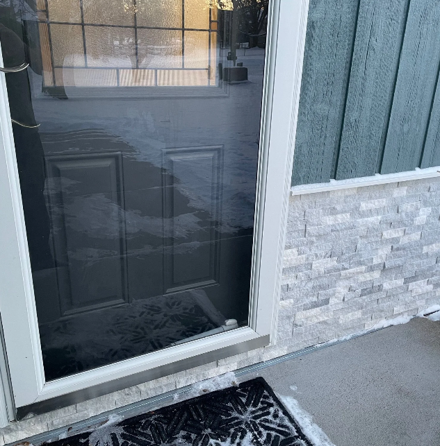 Close-up of a glass door with a black and white patterned doormat outside, next to a blue-green wooden wall with stone veneer at the bottom.
