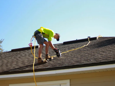 A worker on a ladder cleaning or inspecting a roof, wearing a yellow safety vest, shorts, work boots, and gloves, with a safety harness attached to a rope.