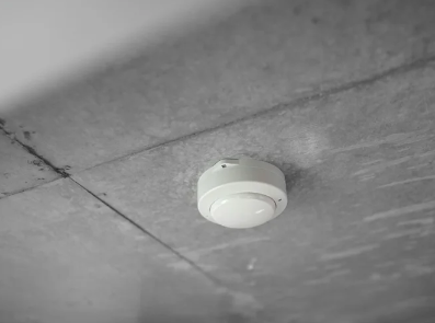 A white smoke detector mounted on a gray tiled ceiling.