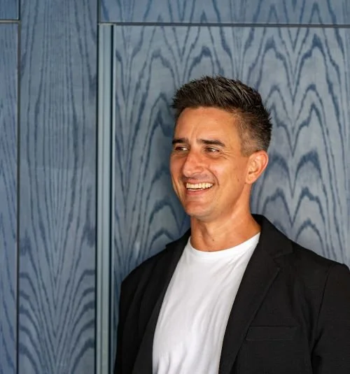 A man smiling, wearing a white t-shirt and black blazer, standing against a blue wooden wall.